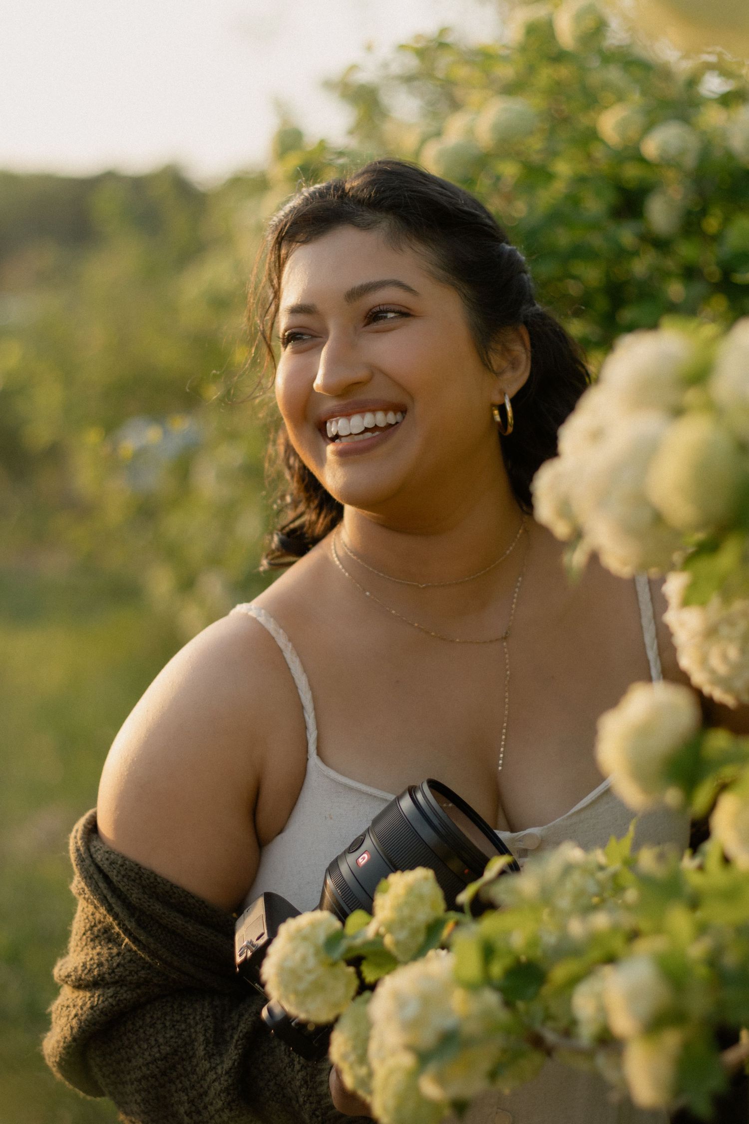 A joyful portrait in golden sunlight surrounded by blooming white roses and wearing a brown cardigan over white top.