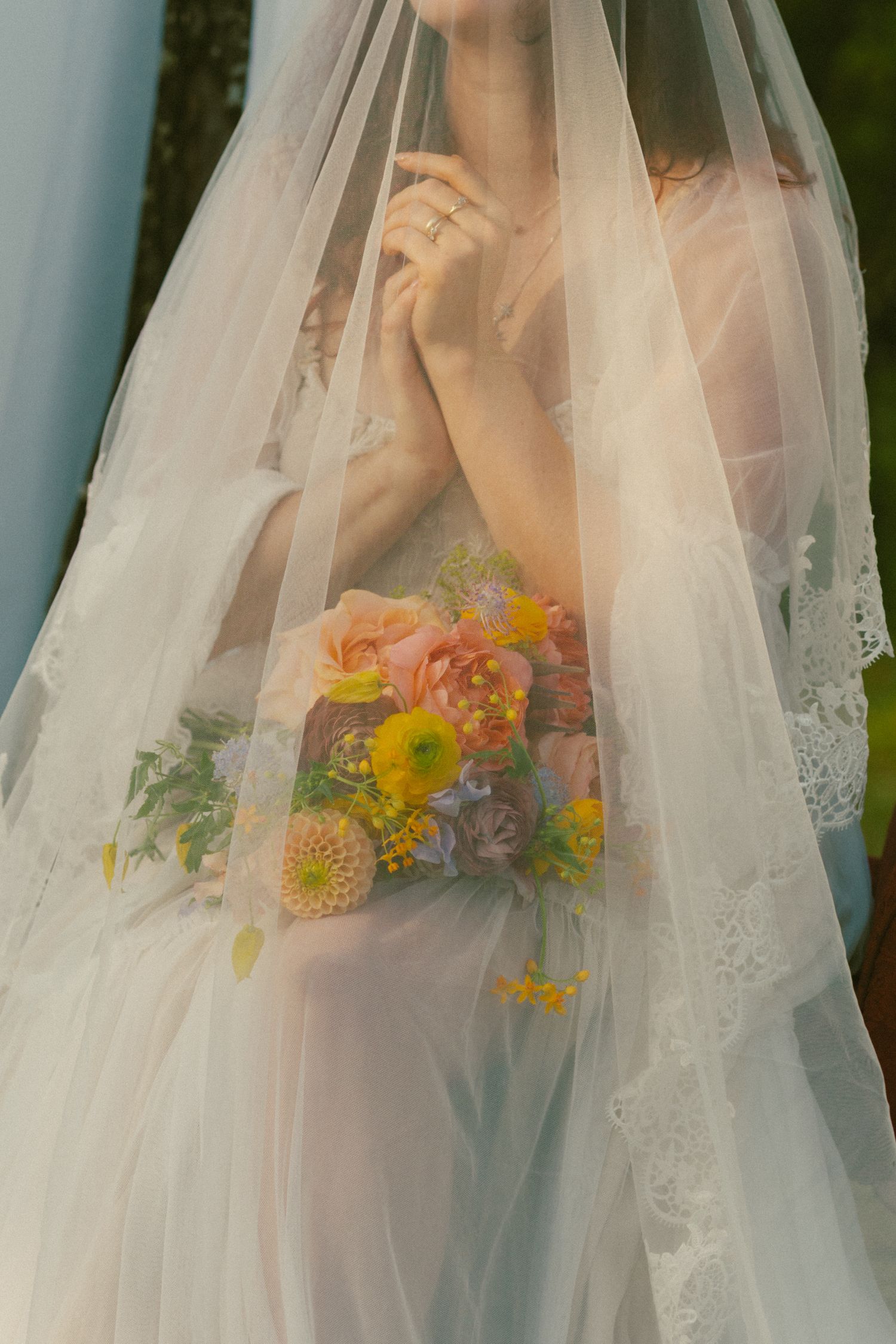 A delicate lace wedding veil frames a vibrant sunflower and wildflower bouquet against a soft background.