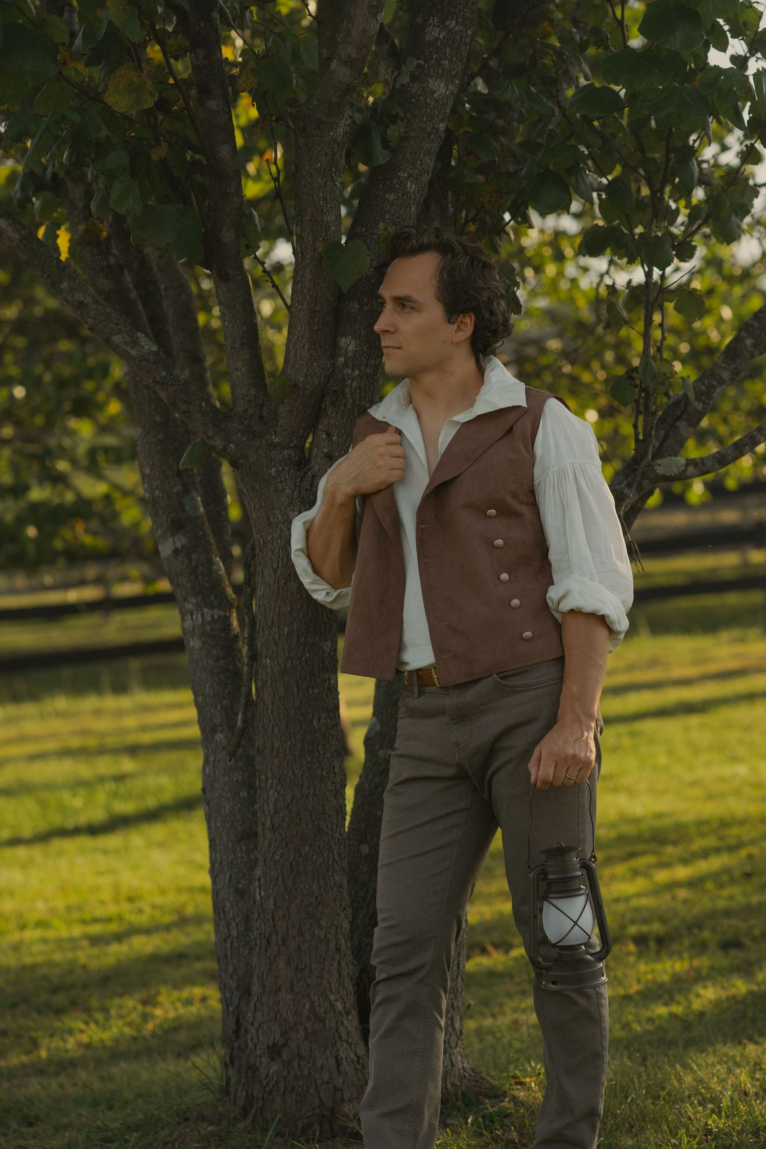 A person in period clothing leans against a tree in a sunlit orchard wearing a brown vest and white shirt.
