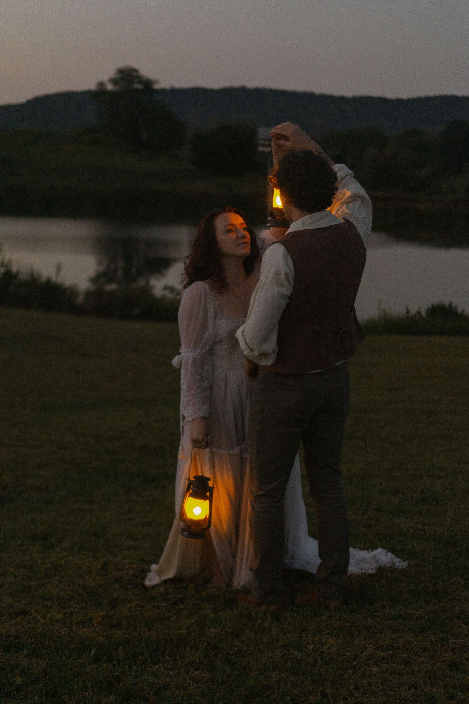 A couple shares a romantic moment at dusk by a lake while holding a glowing lantern in a dreamy outdoor setting.