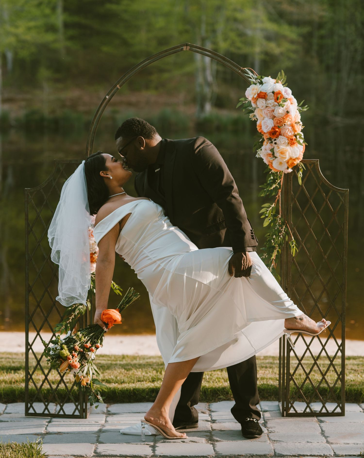 A couple in wedding attire playfully poses on a wooden deck near a floral-decorated trellis by a lake.