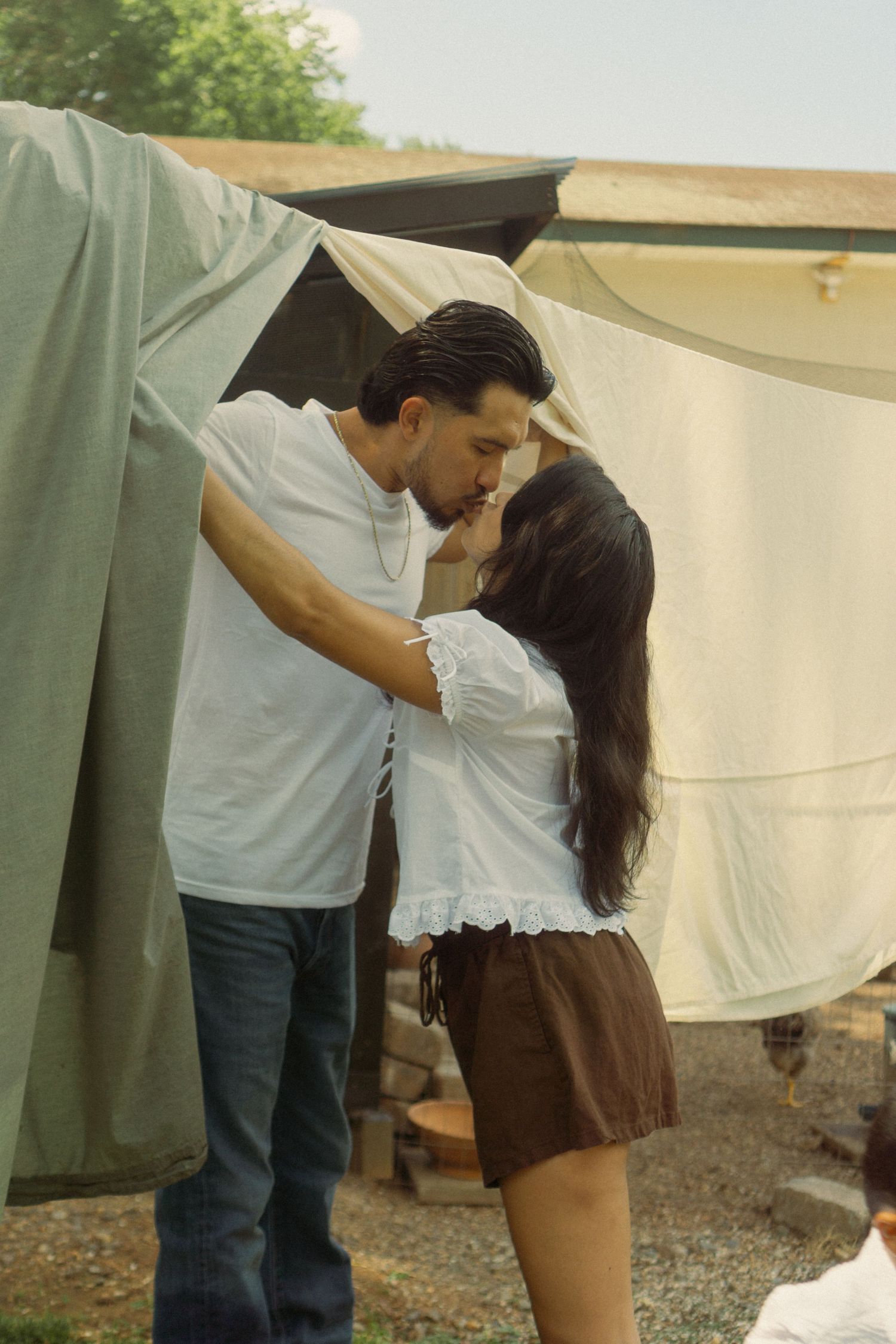 A couple interacts near white hanging fabric in an outdoor setting.