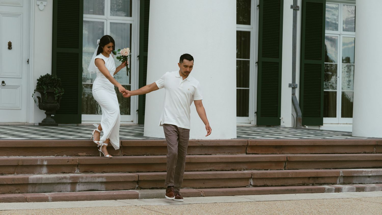 A couple descends white stairs outside a classic building in formal attire.