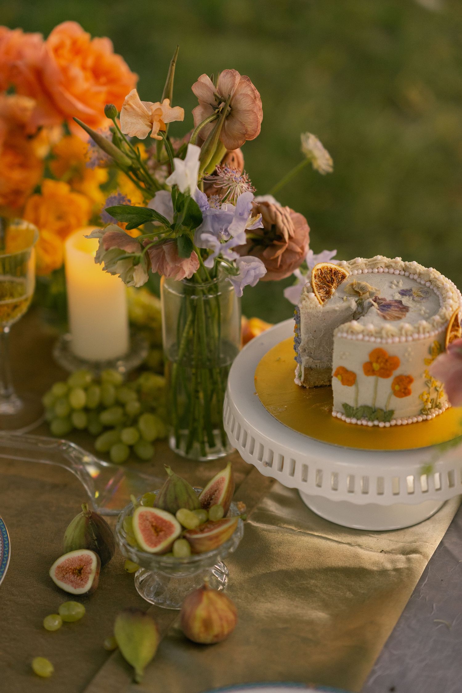 Elegant table setting with orange flowers, figs, and a decorative white cake stand adorned with fresh fruit.