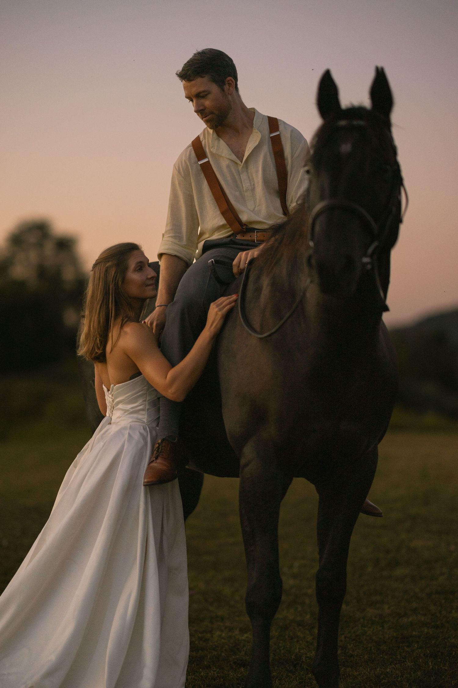 A bride in white gown stands beside a dark horse at sunset, gazing up at a man in suspenders seated on horseback.