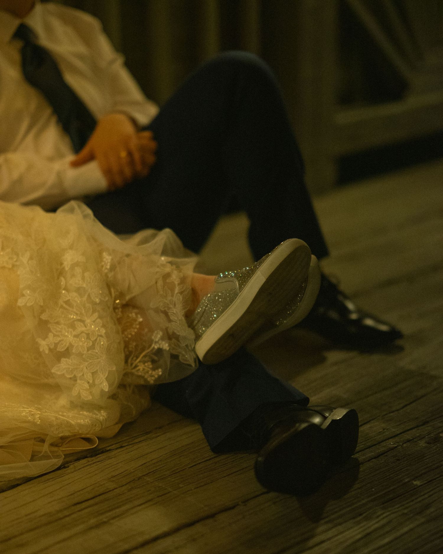 Close-up of formal dress shoes and wedding gown hem on wooden floorboards in warm lighting.