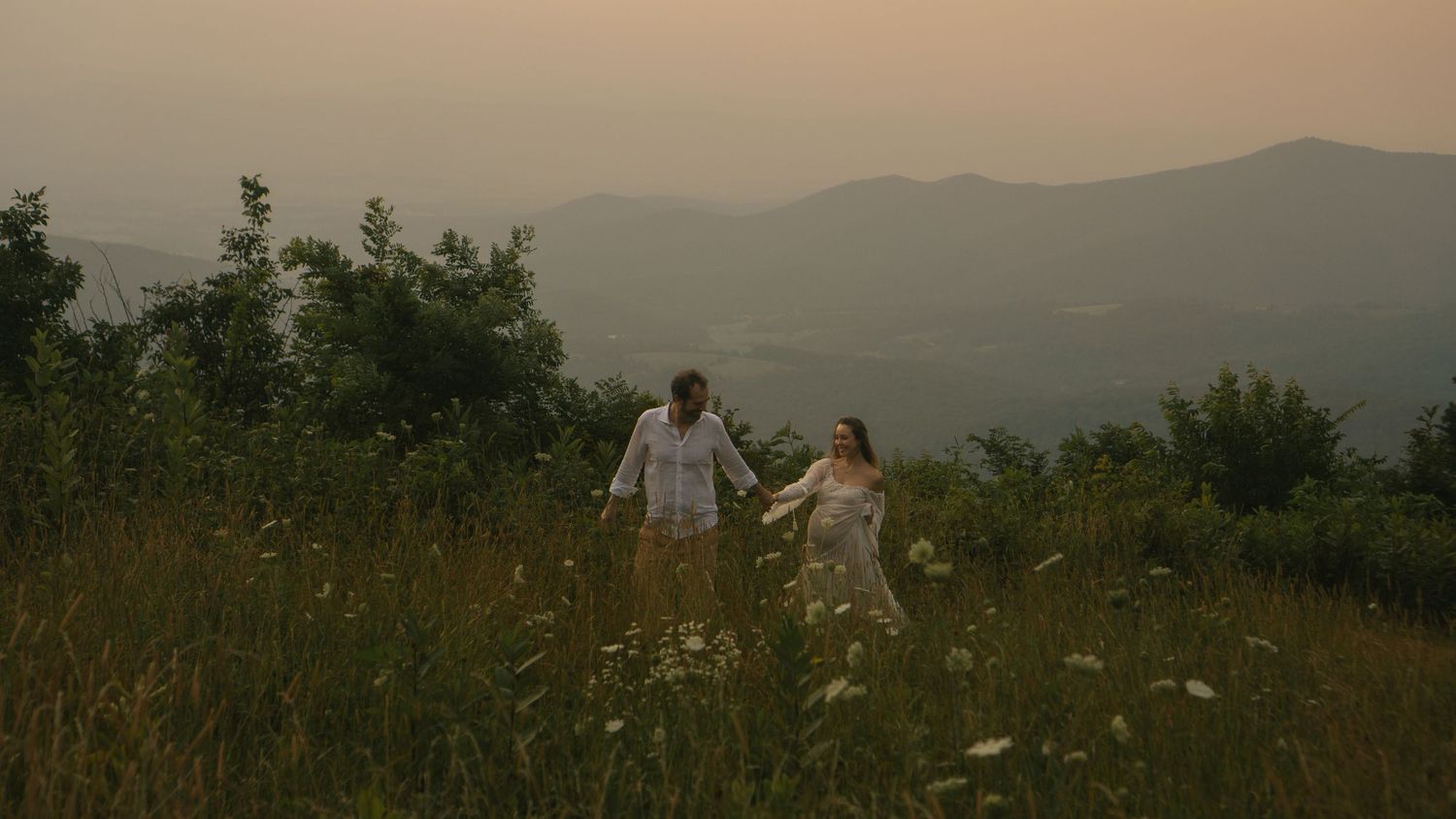 A couple walking through a field of tall grass and wildflowers, a beautiful view of the Shenandoah Mountains are behind them.