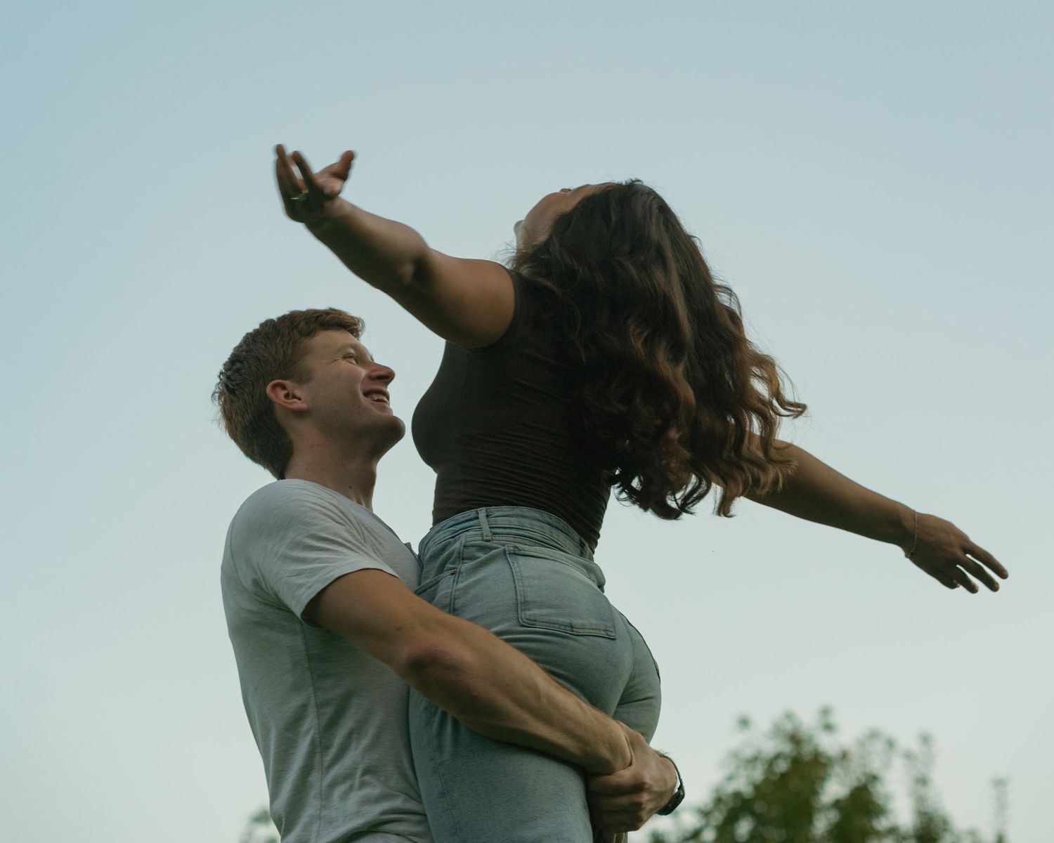 a young man lifting his fiancé up in the air while she throws her head and arms back, facing up towards the clear, blue hour sky. Vint Hill
