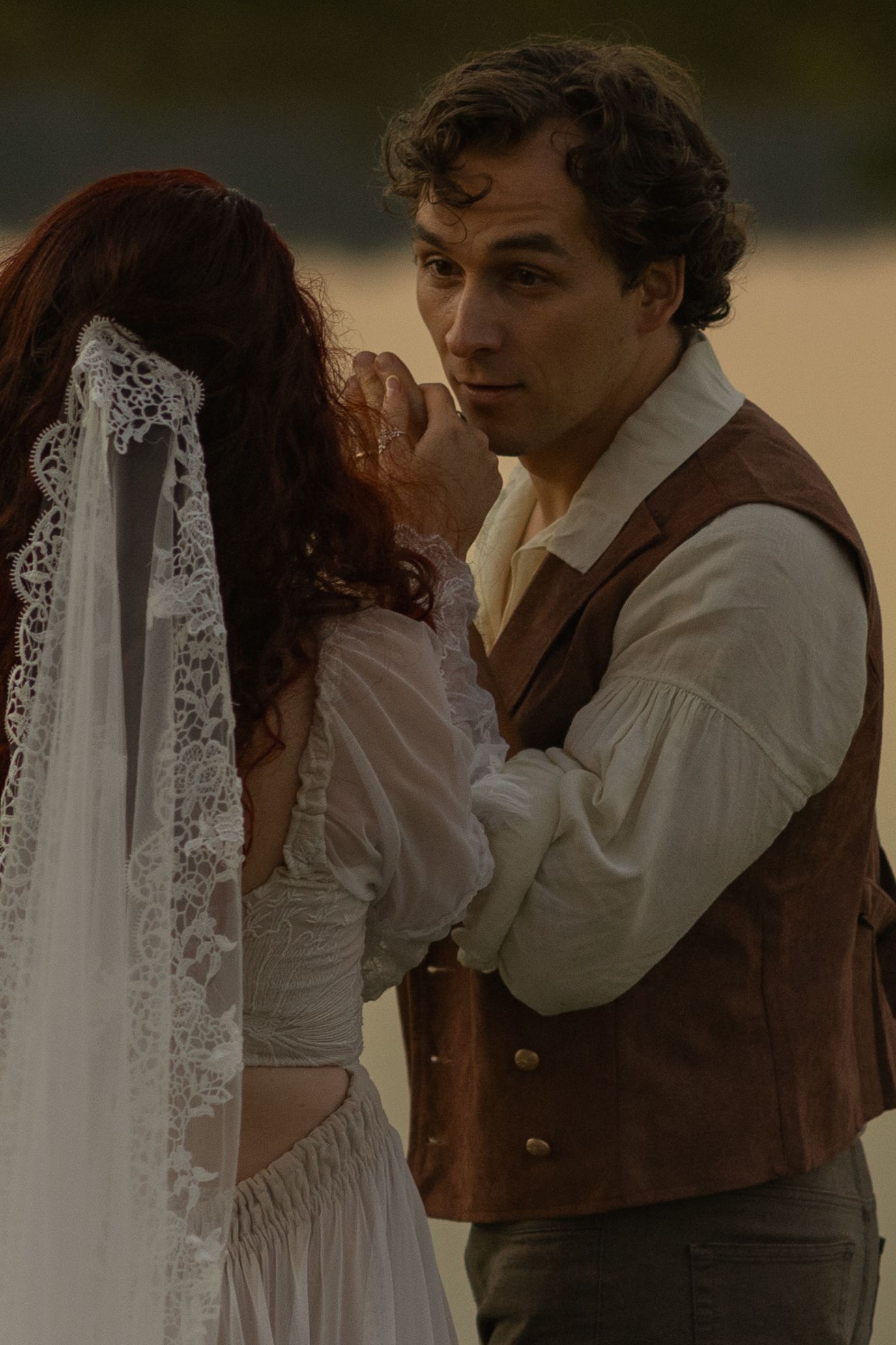 A romantic couple in period clothing embrace in warm sepia tones as white lace curtains flow in the background.