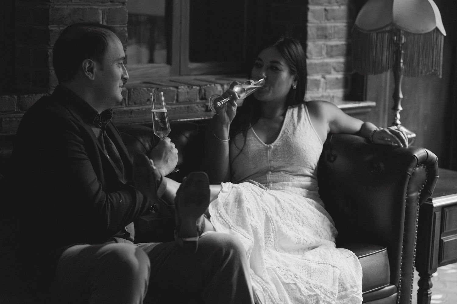 Black and white image of newlyweds sharing drinks on vintage furniture.
