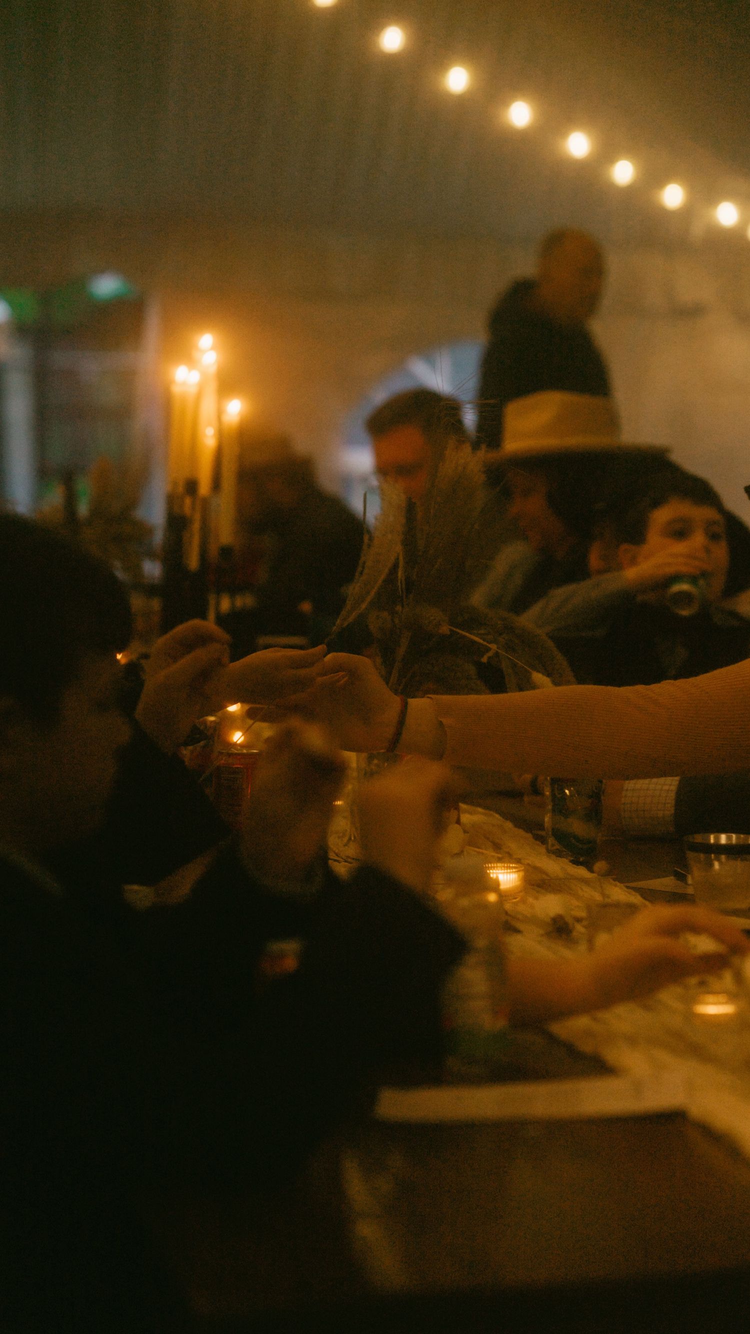 Guests clink glasses at a dimly lit dinner party with warm string lights and candles creating an intimate ambiance.