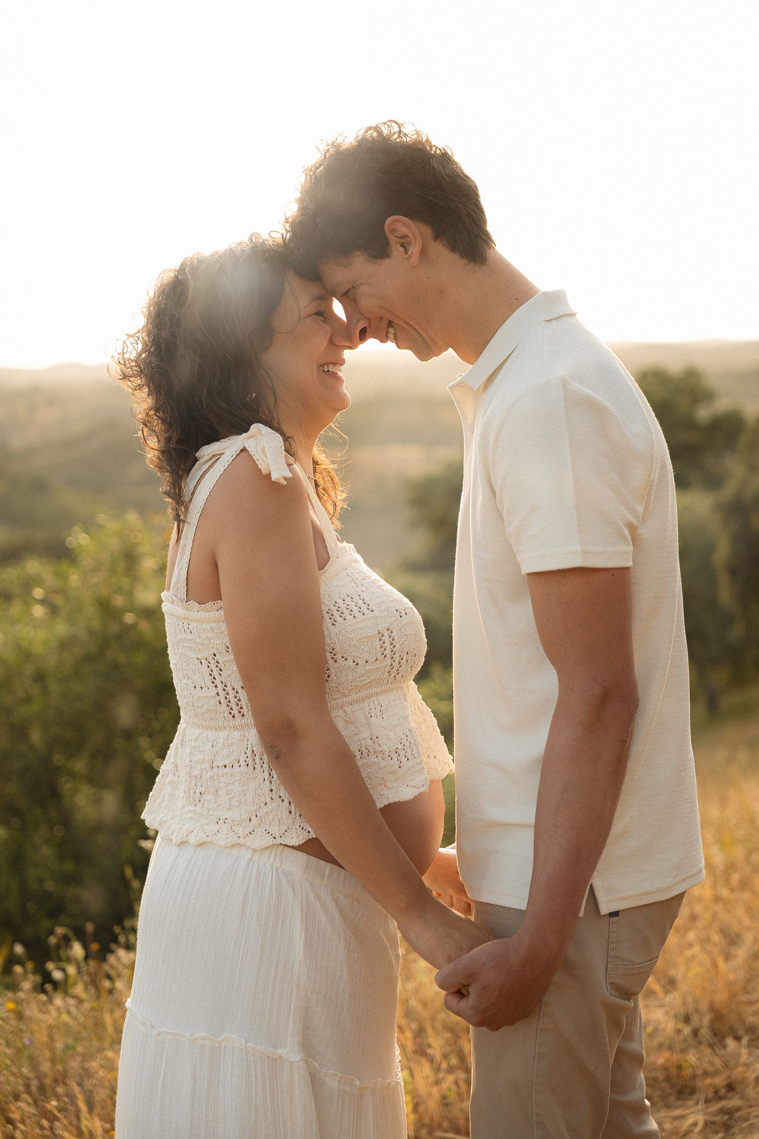 Romantic couple holding hands and touching foreheads at golden sunset in a field, wearing white outfits.