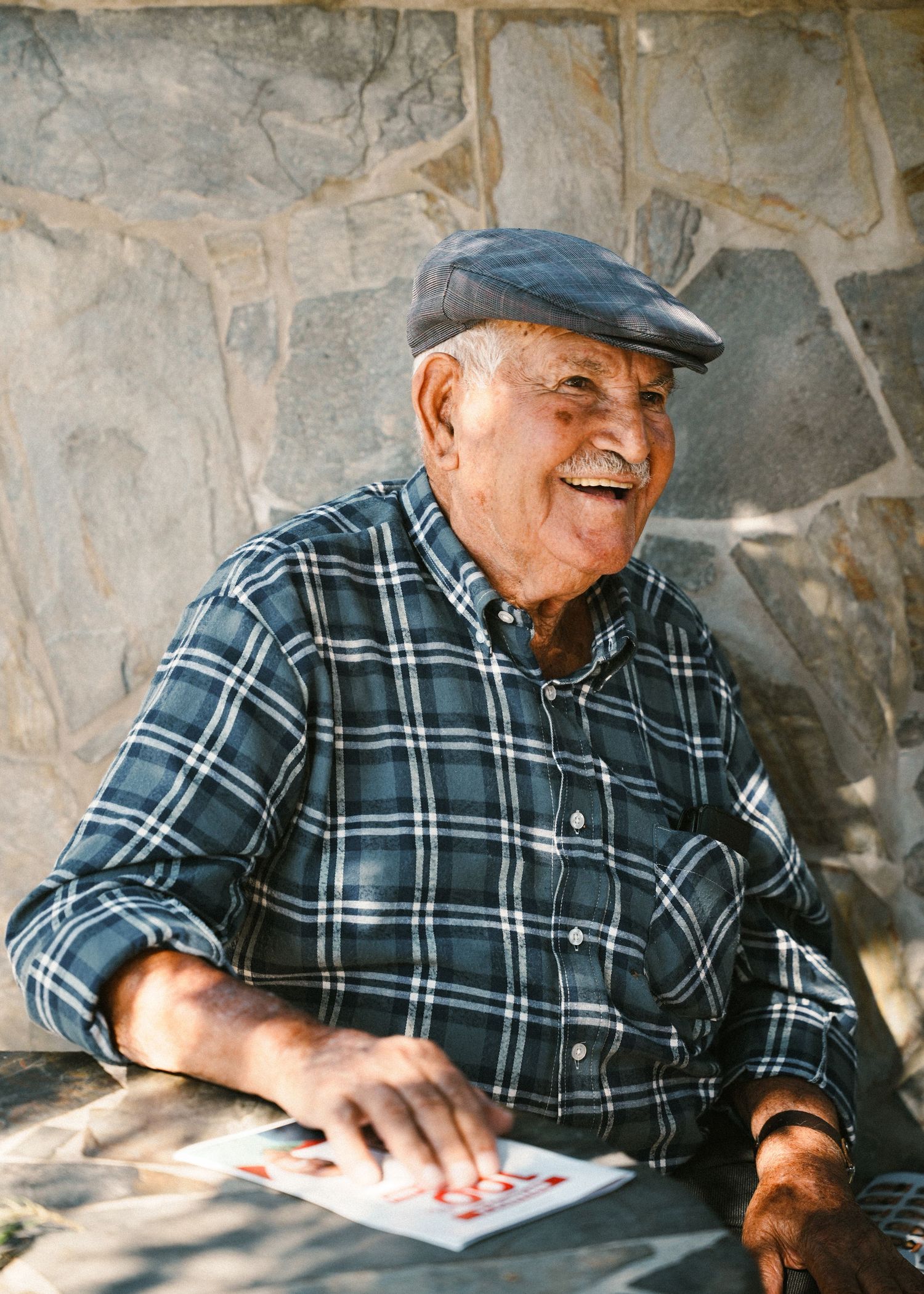 Senior in plaid shirt and cap playing cards by stone wall.