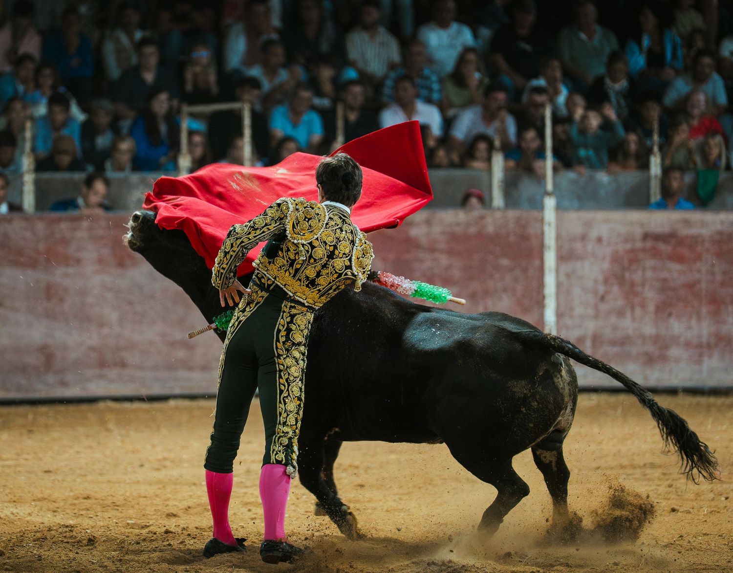 Matador in traditional gold embroidered suit waves red cape during dramatic bullfight in sandy arena.