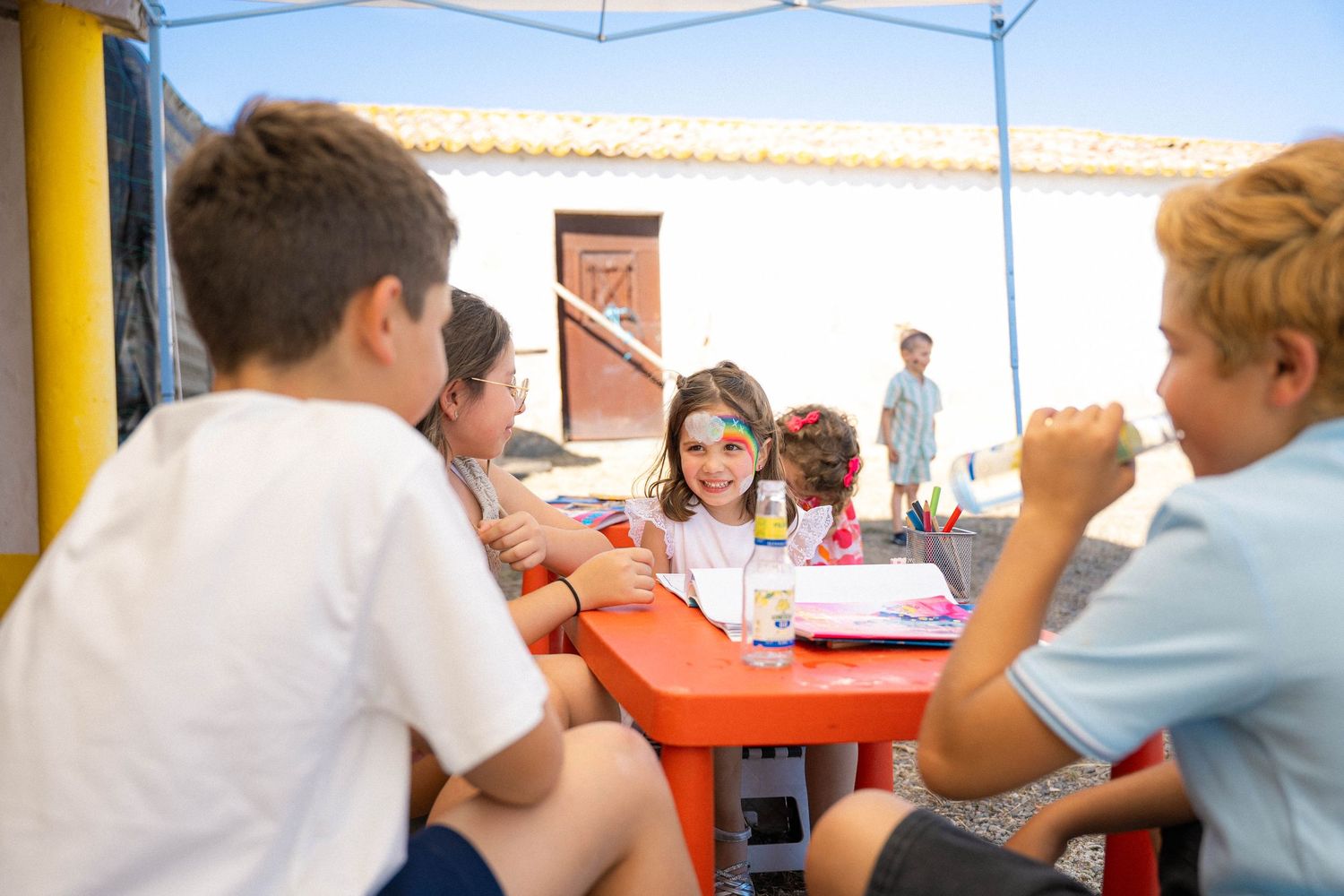 Children sitting around a red table outdoors under a blue canopy, smiling and enjoying time together.