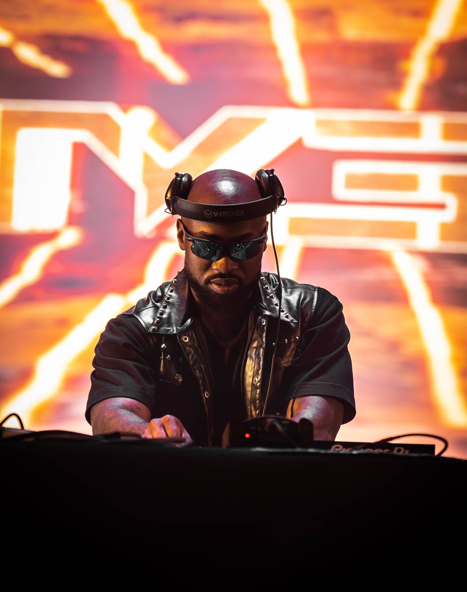 DJ performs at an electronic music event with dramatic orange lighting and visual effects in the background.