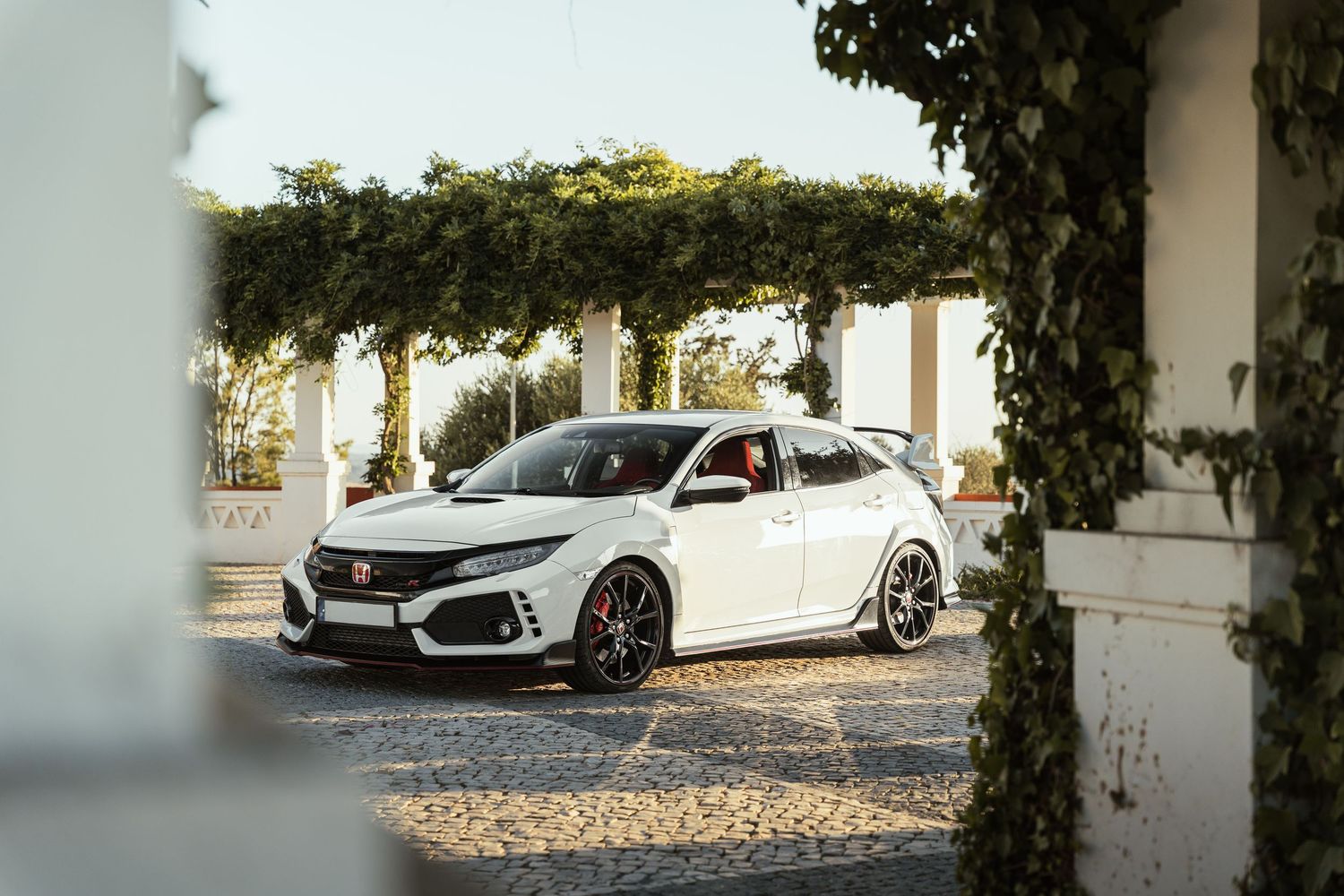 White Honda Civic Type R parked between white pillars with green foliage.