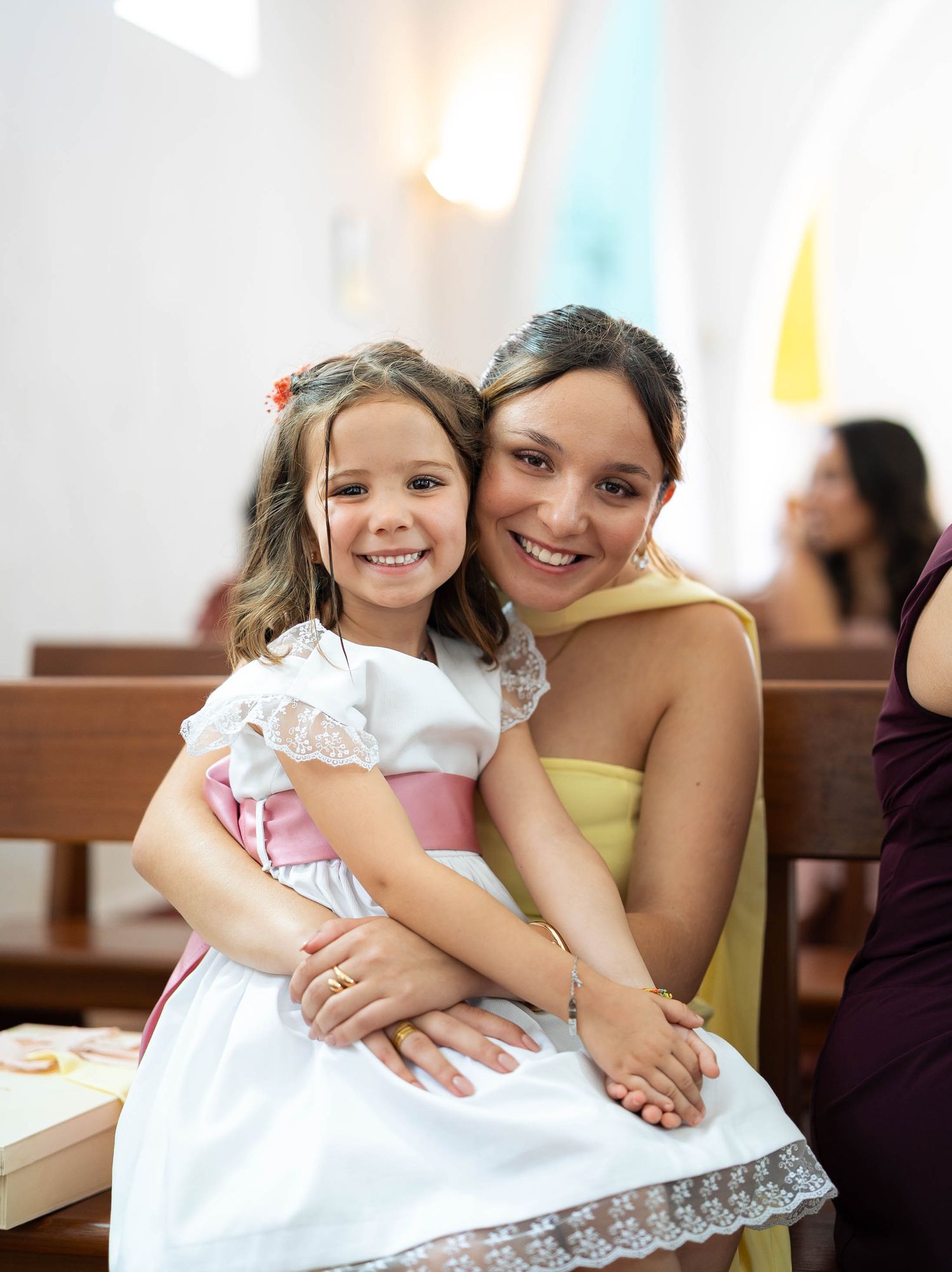 A woman in a yellow dress smiles with a young girl in a white and pink dress inside a bright church.