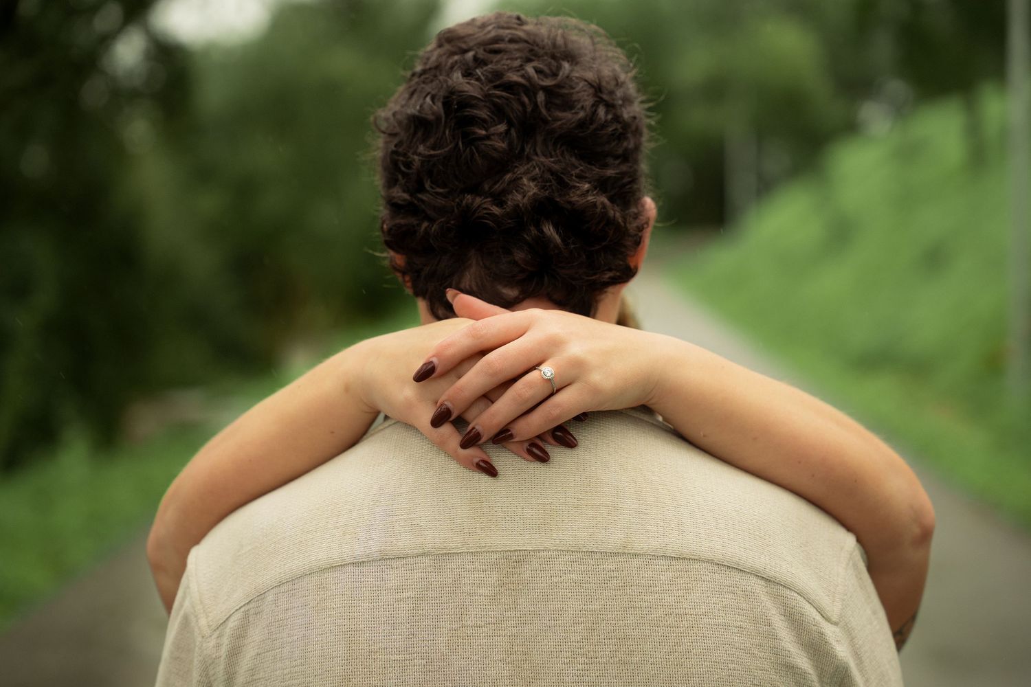 Person seen from behind with curly hair, wearing a beige shirt, with a hand resting on their shoulder outdoors.