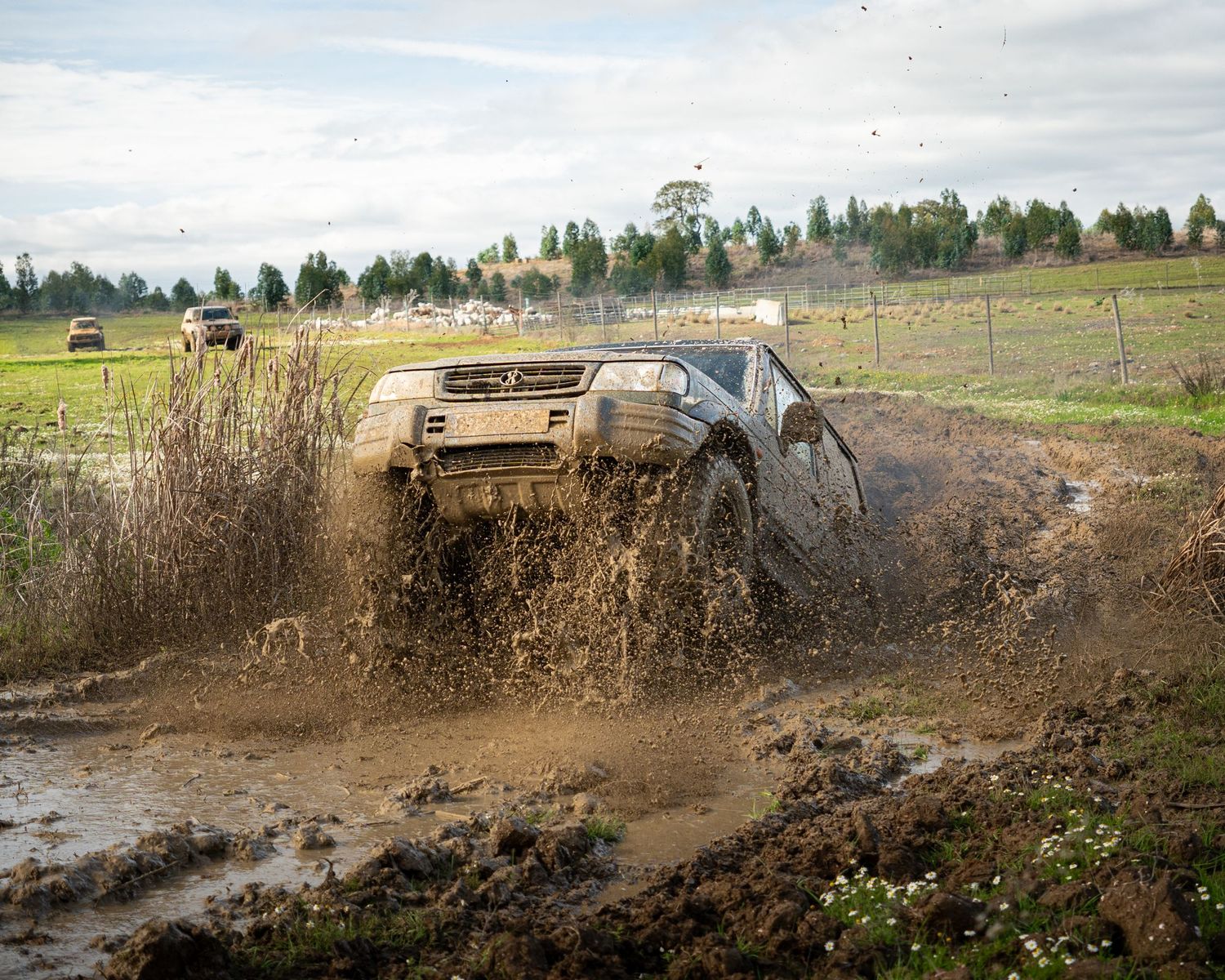 A black SUV kicks up mud and water while off-roading through a muddy terrain on a cloudy day.