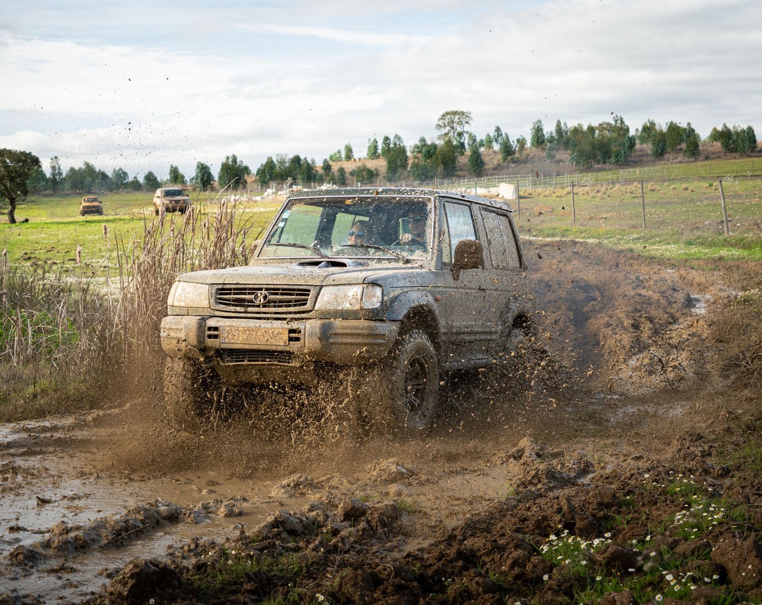 A silver pickup truck splashes through deep mud on an off-road trail, kicking up dirt and debris in a rural setting.