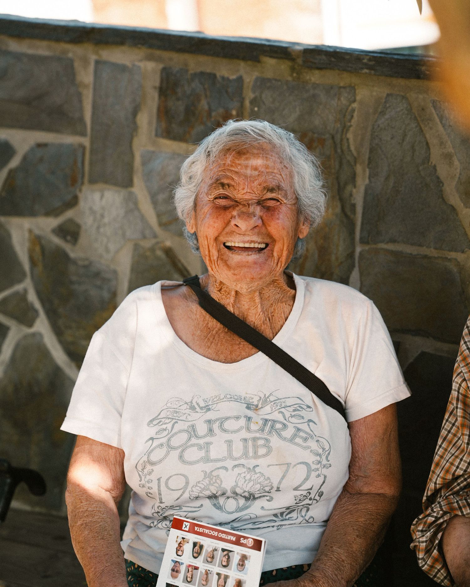 Senior in white t-shirt smiling brightly against stone wall.