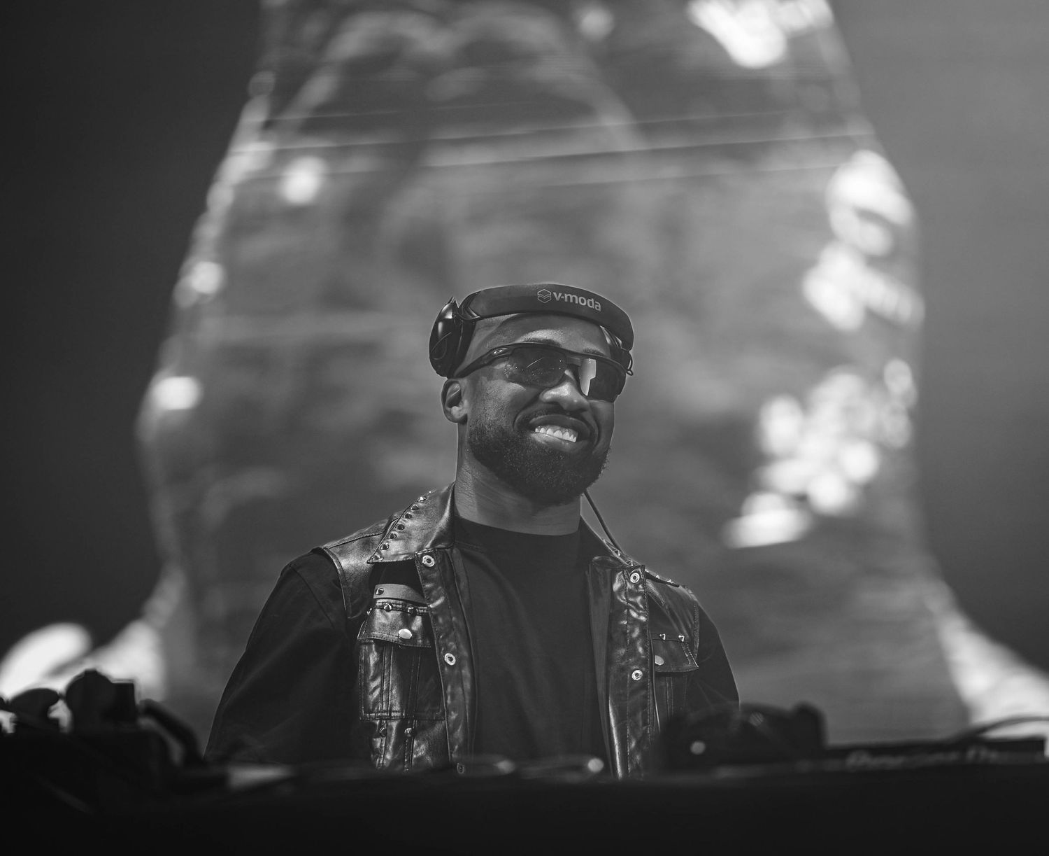 Black and white photo of a DJ performing behind decks at a nightclub with moody lighting.