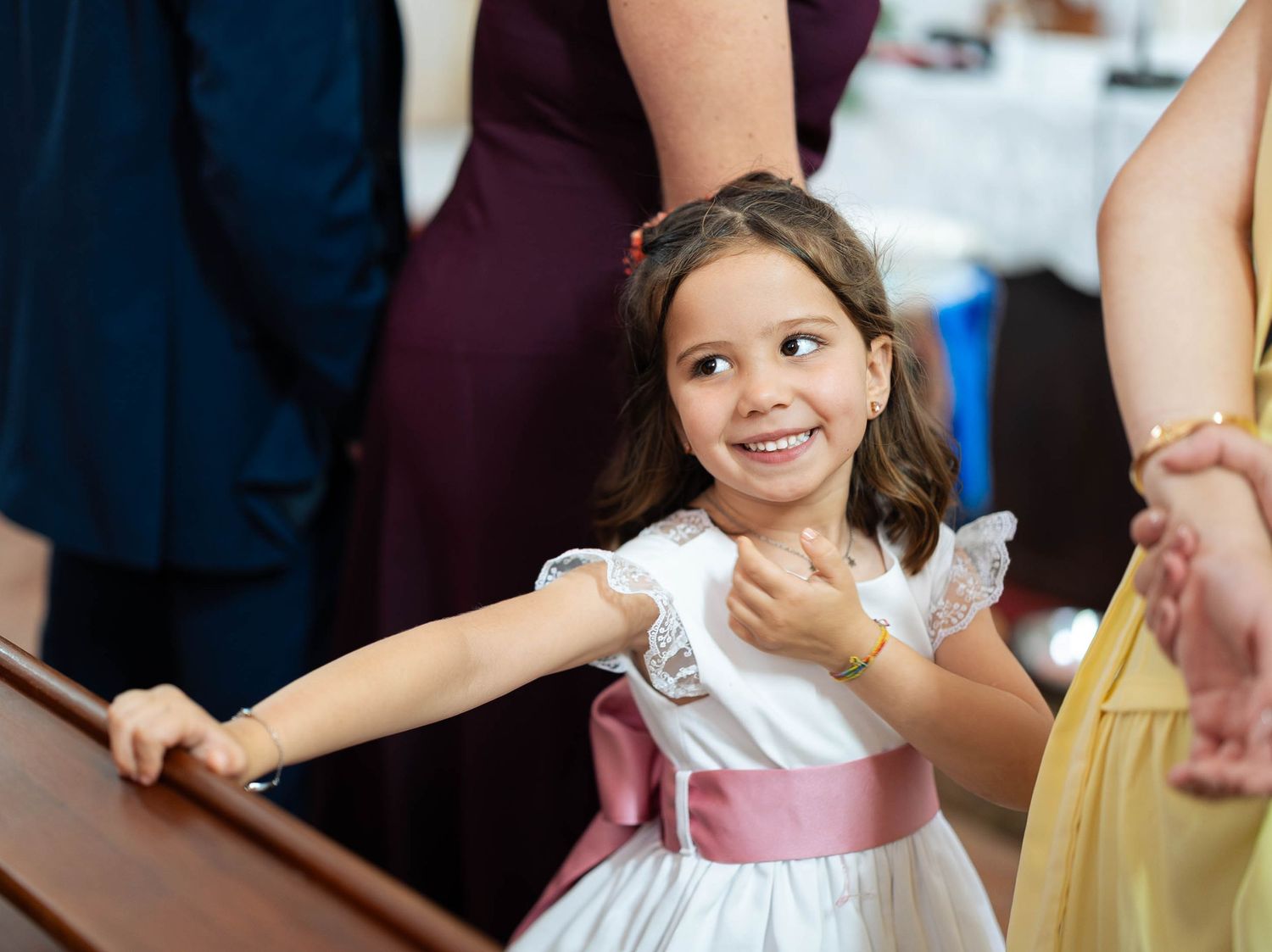 Young flower girl in white dress with pink sash smiles joyfully at a wedding ceremony.