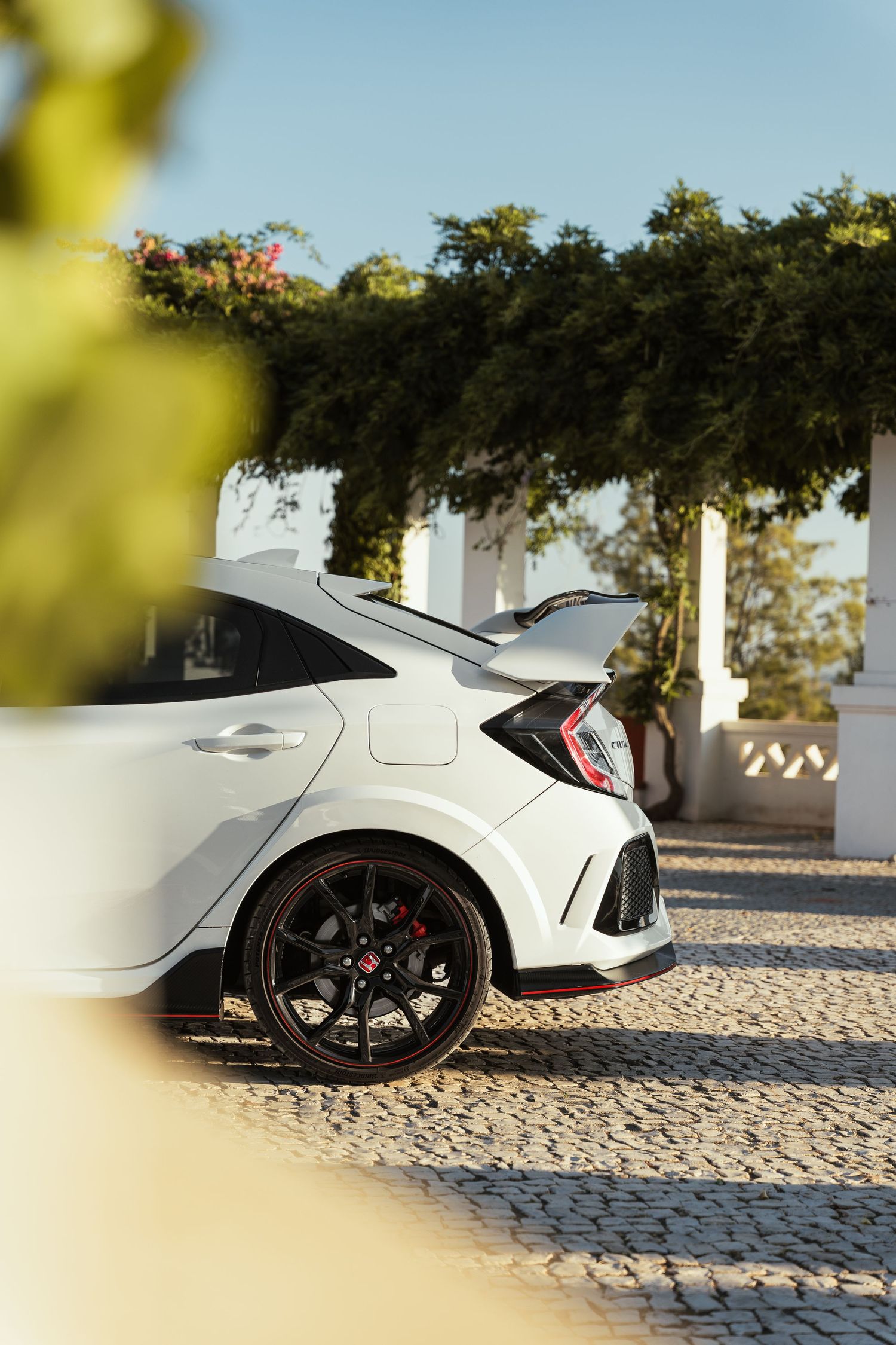 Side profile view of a white Honda Civic Type R on cobblestone ground with trees in background.