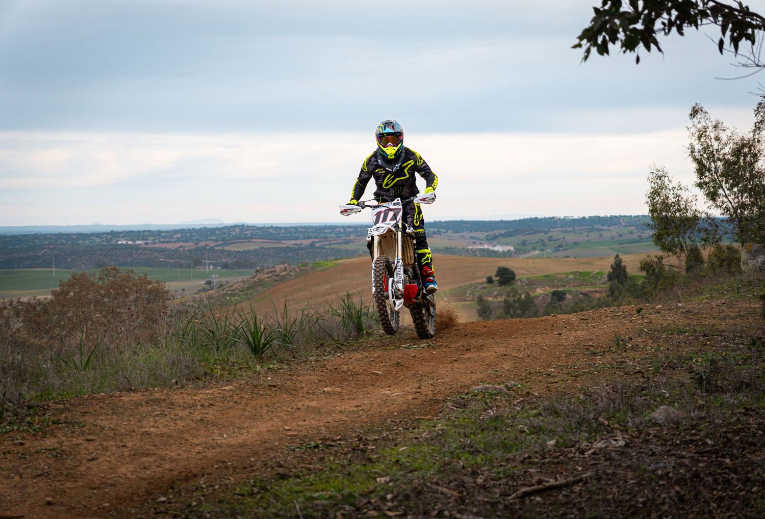 Dirt bike riders traverse scenic mountain trails with rolling hills and valleys in the background.