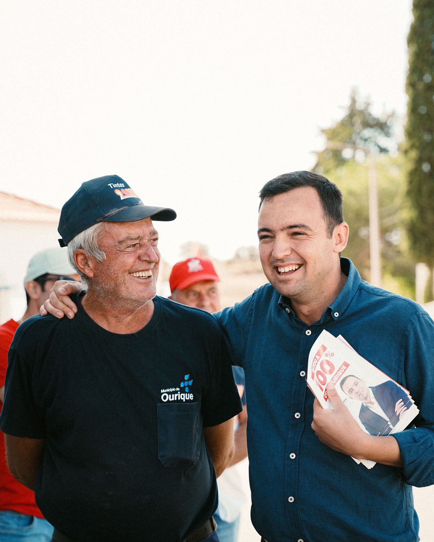 Two people sharing a laugh while holding campaign materials outdoors.