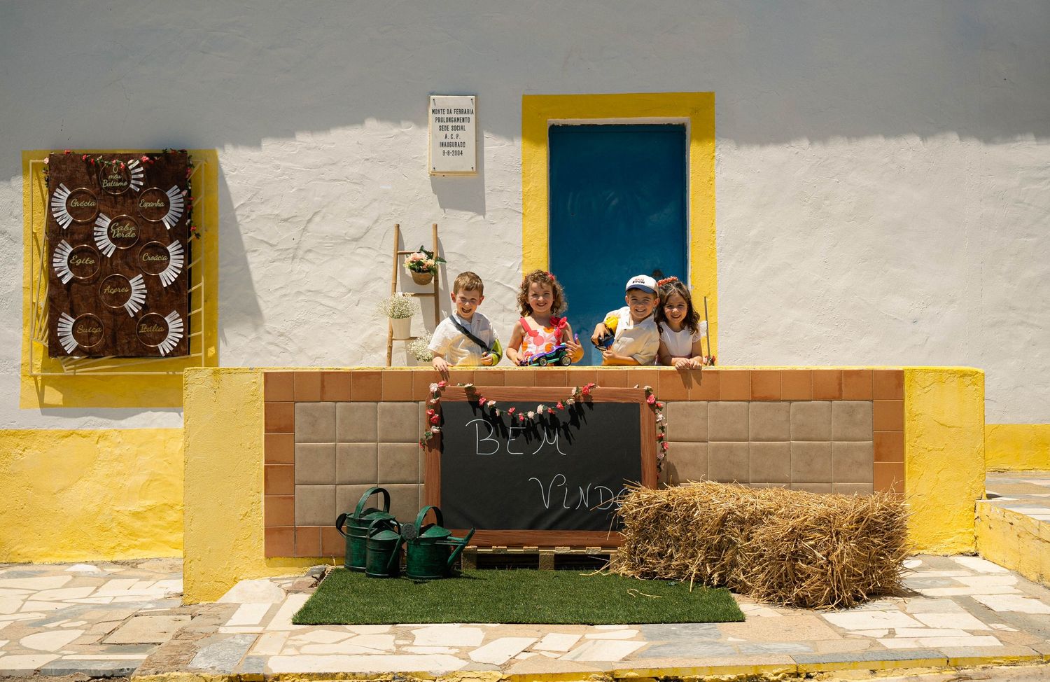 Children playing behind a yellow-trimmed wall with a chalkboard sign and hay bale outside a white building.