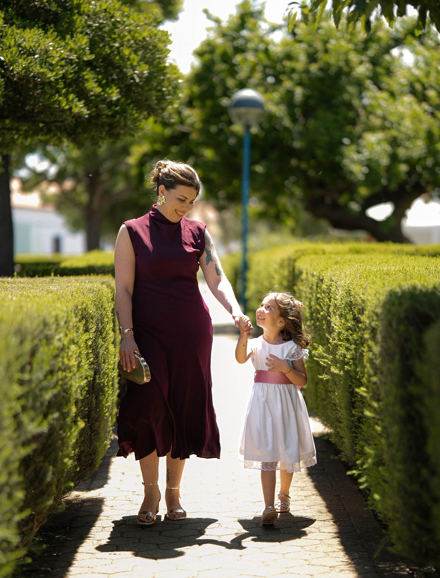 A woman in a burgundy dress holds hands with a little girl in white as they walk through a sunny garden path.