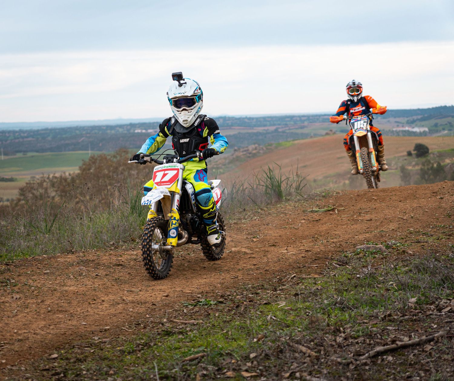 Dirt bike riders navigate a winding off-road trail through green hills and rugged terrain during an outdoor motorsport event.