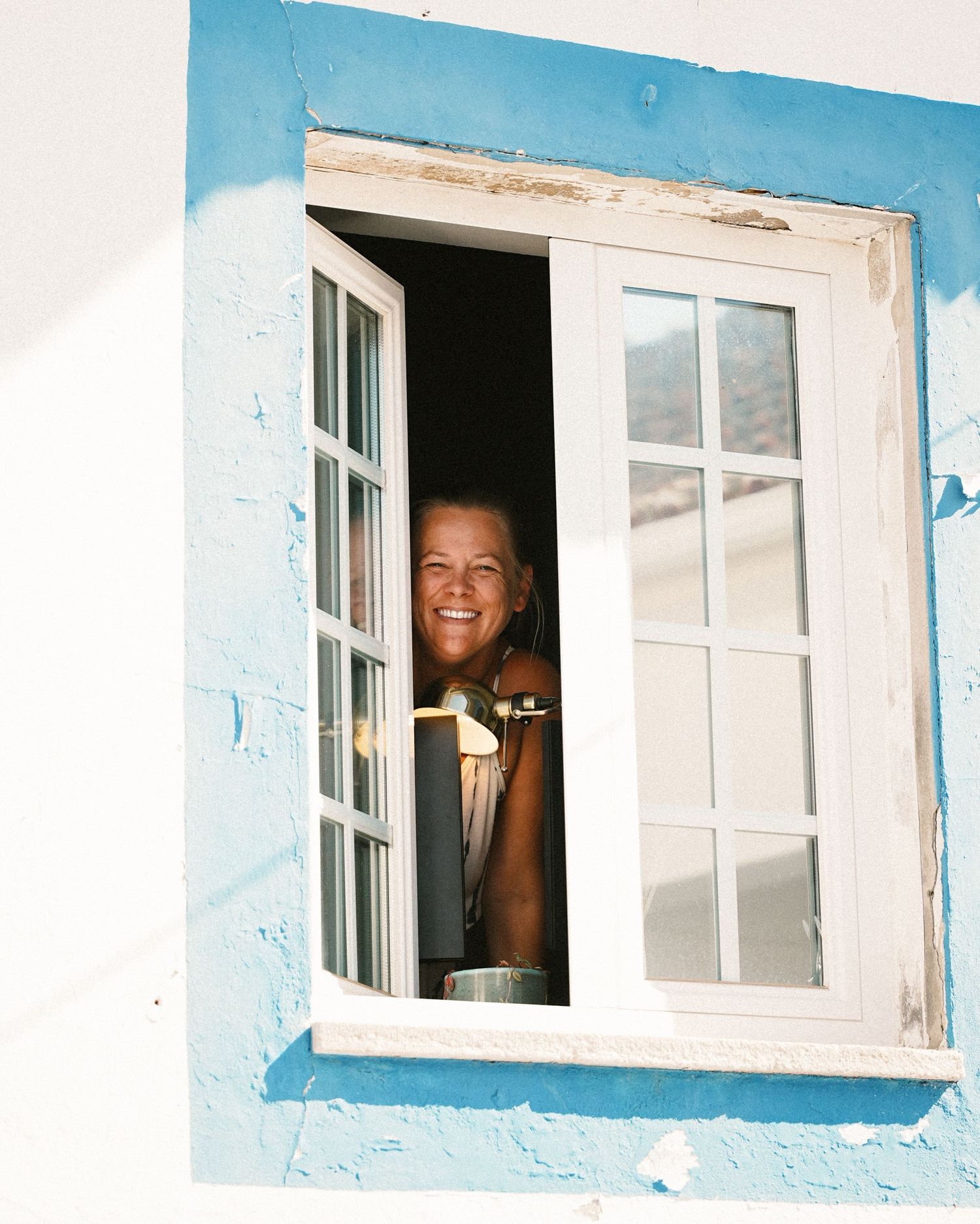 Someone smiling and waving through a white-framed window of a blue building.