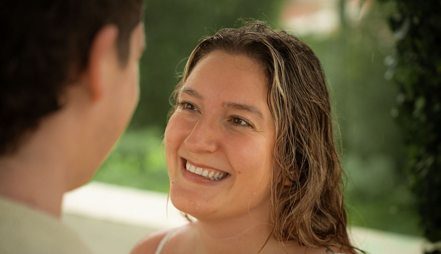 A smiling woman with wet hair looks up at someone outdoors in warm sunlight.