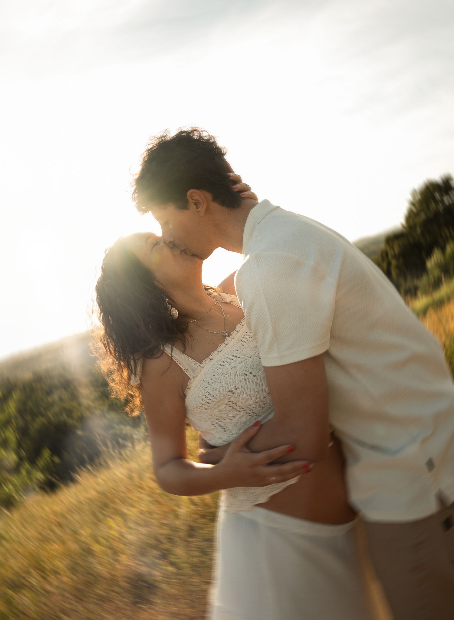 A couple shares a romantic kiss in a sunlit golden field, the woman in a white lace dress embraced by her partner.