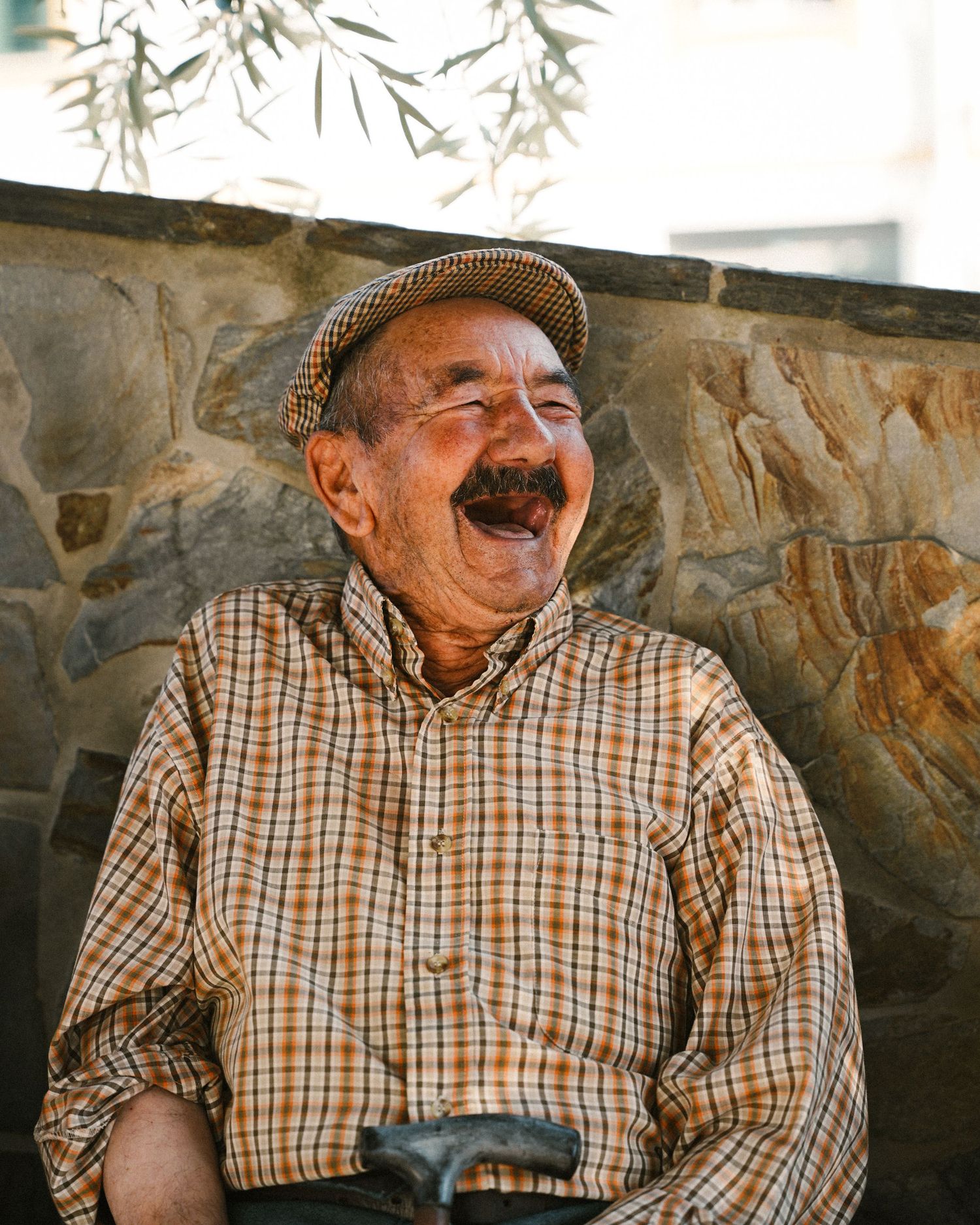 Elderly person in plaid shirt and flat cap smiling against stone wall background.