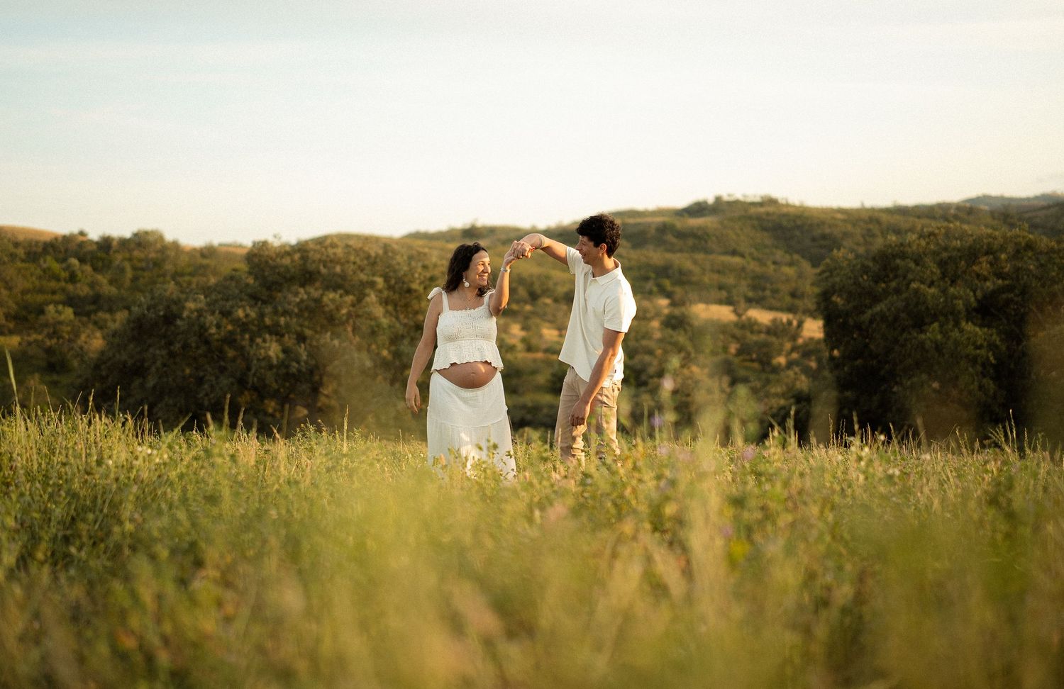 Pregnant couple holding hands in a sunlit green meadow with rolling hills and trees in the background.