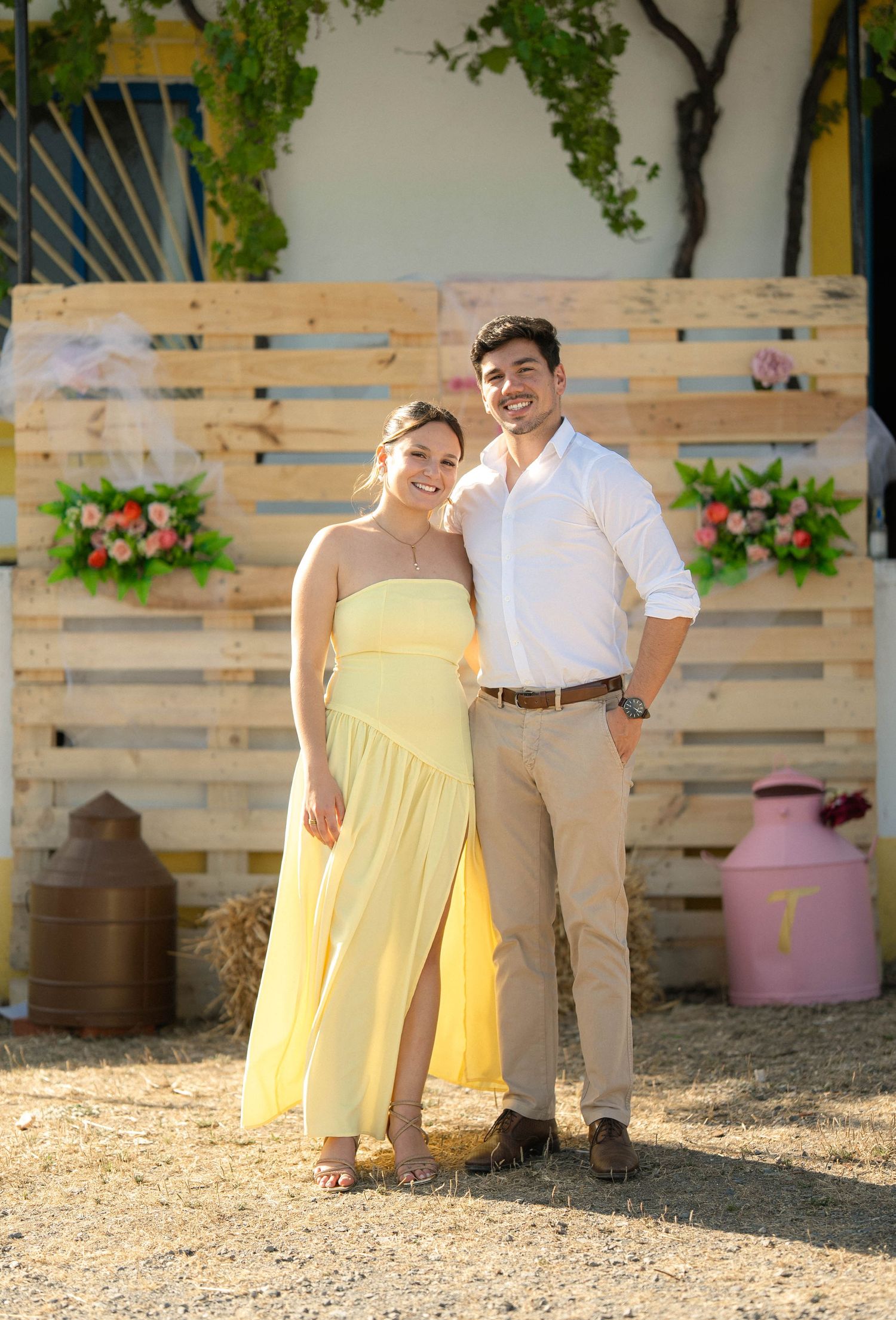 Couple posing together in front of a rustic wooden pallet backdrop with floral decorations at an outdoor event.