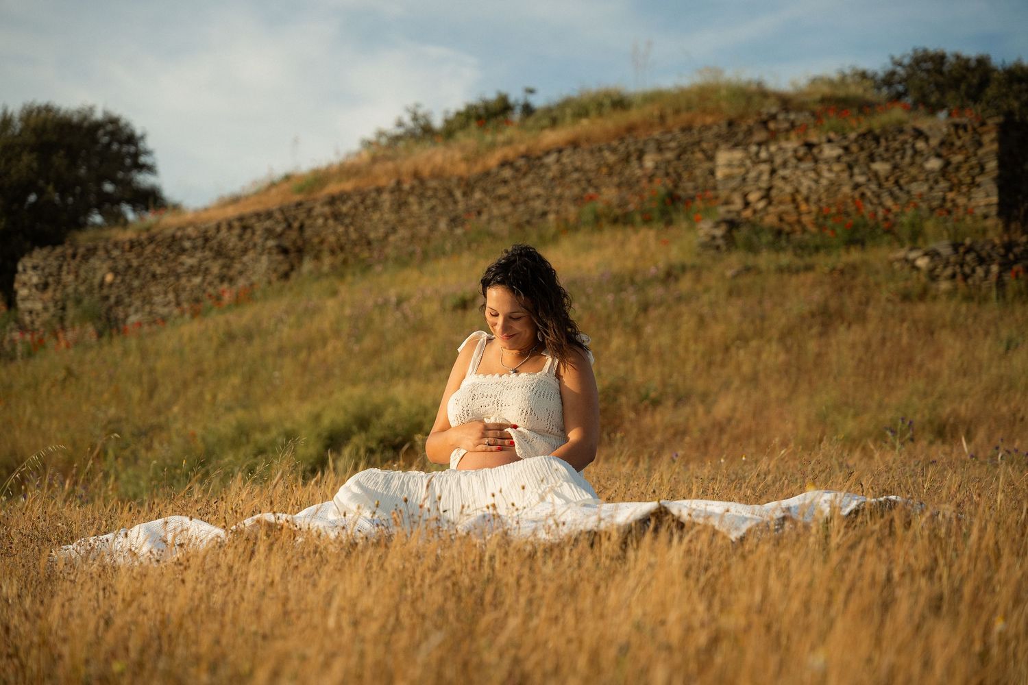 Pregnant woman in white floral dress sitting in golden grass field, cradling baby bump during outdoor maternity photoshoot.