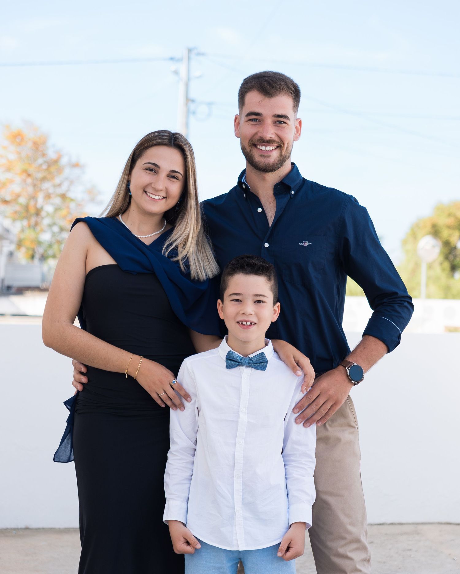Three people in coordinated formal attire posing together outdoors against white wall.