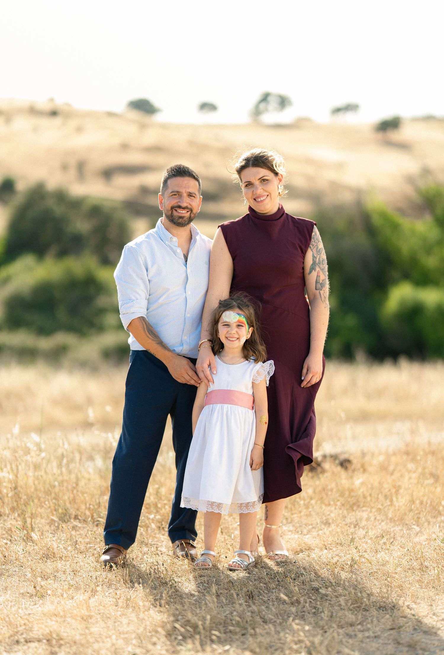 Family of three posing outdoors in golden field, man in white shirt, woman in burgundy dress, young girl in white dress.