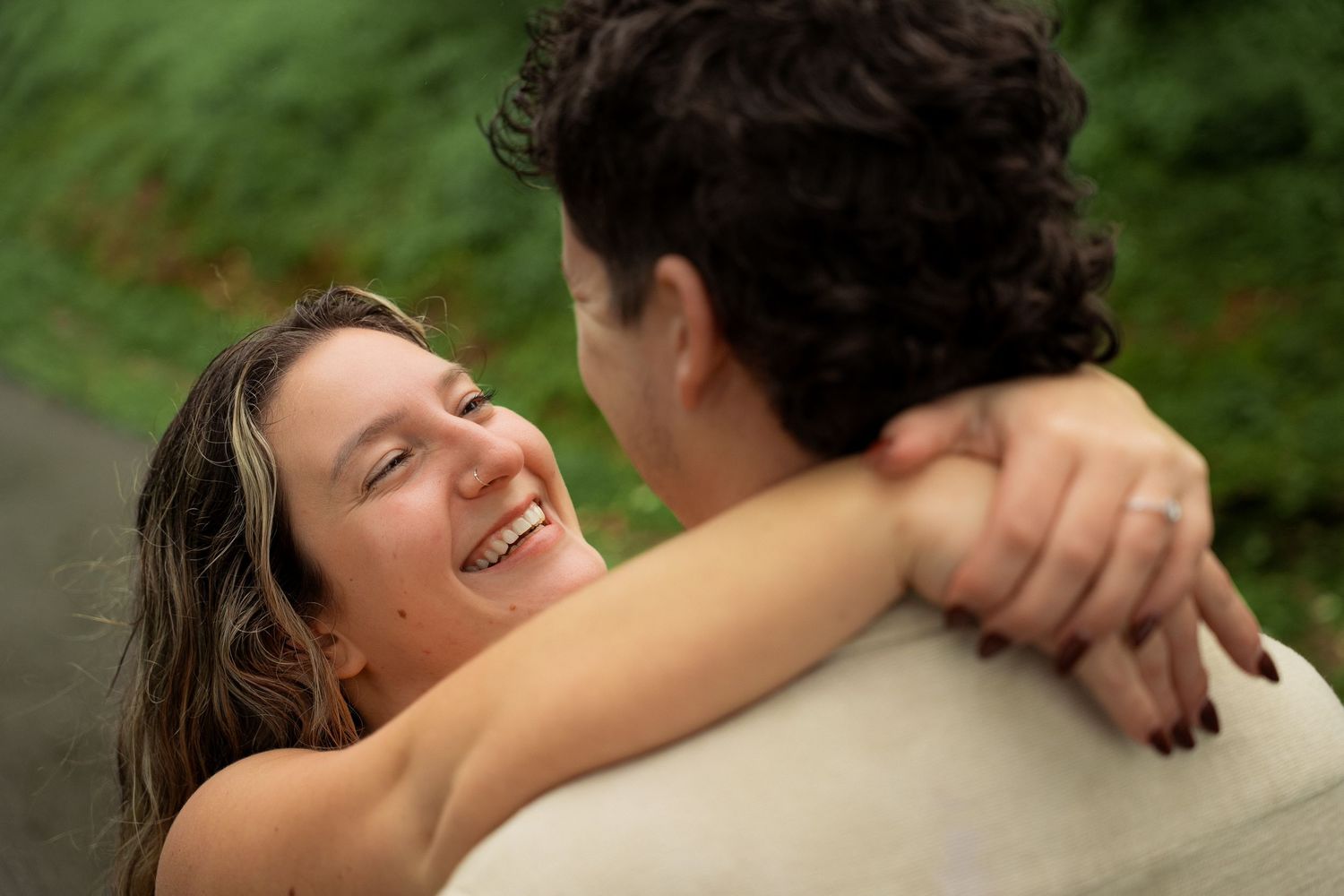 A smiling woman embraces a curly-haired man outdoors, looking up at him joyfully with arms around his shoulders.