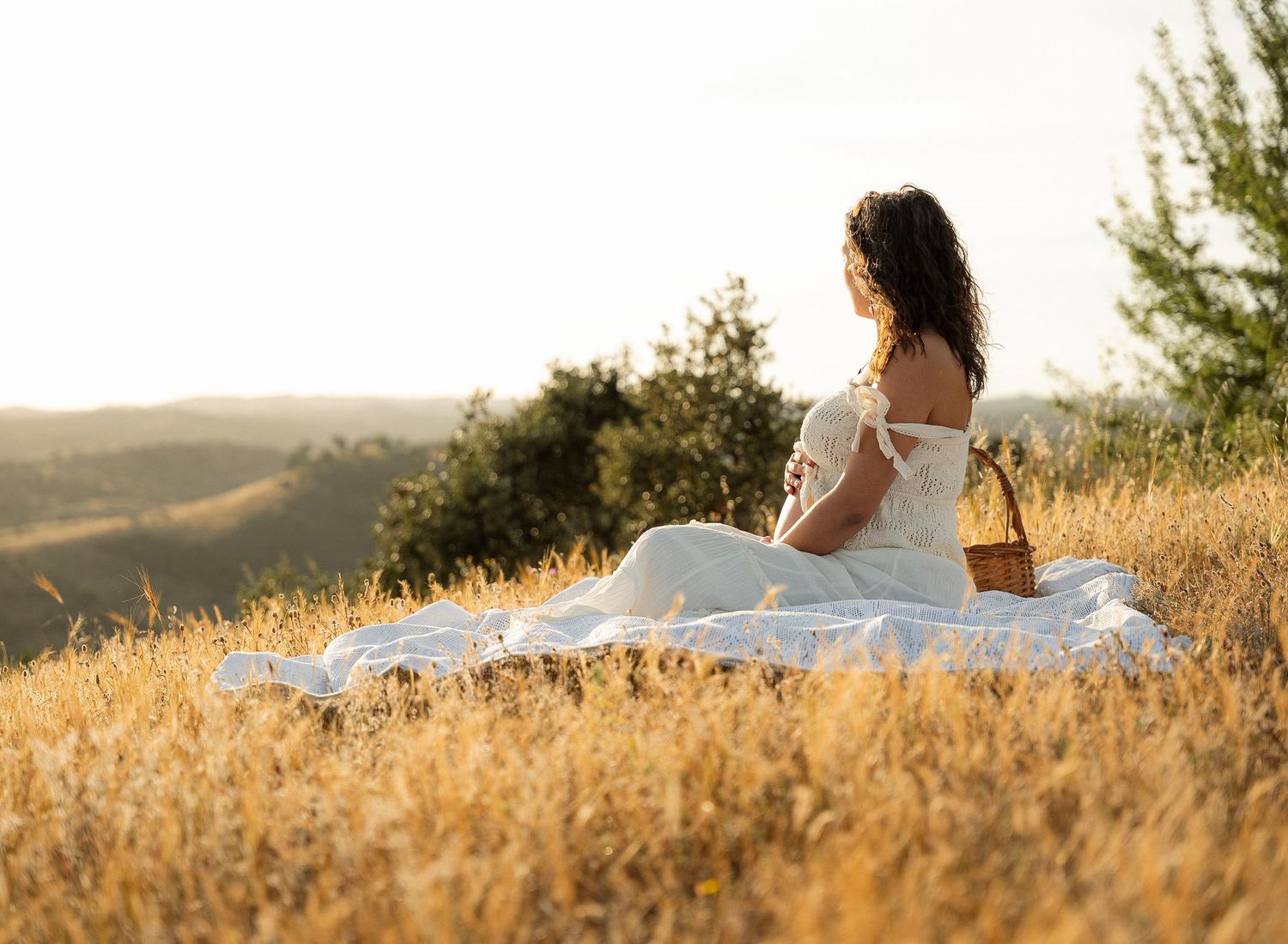 Pregnant woman in white dress sitting on blanket in golden field enjoying scenic countryside view during sunny day.