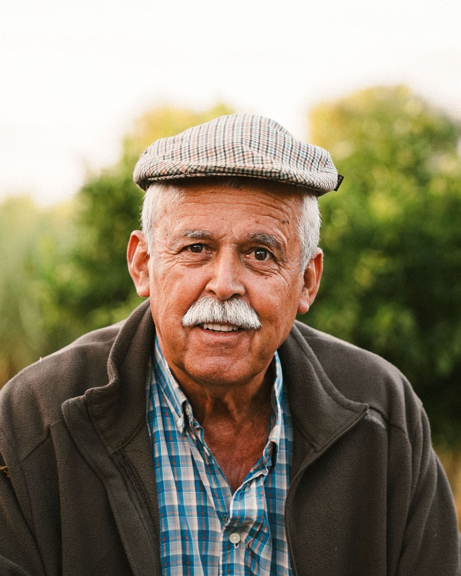 Person wearing a flat cap and plaid shirt sitting outdoors in natural lighting.