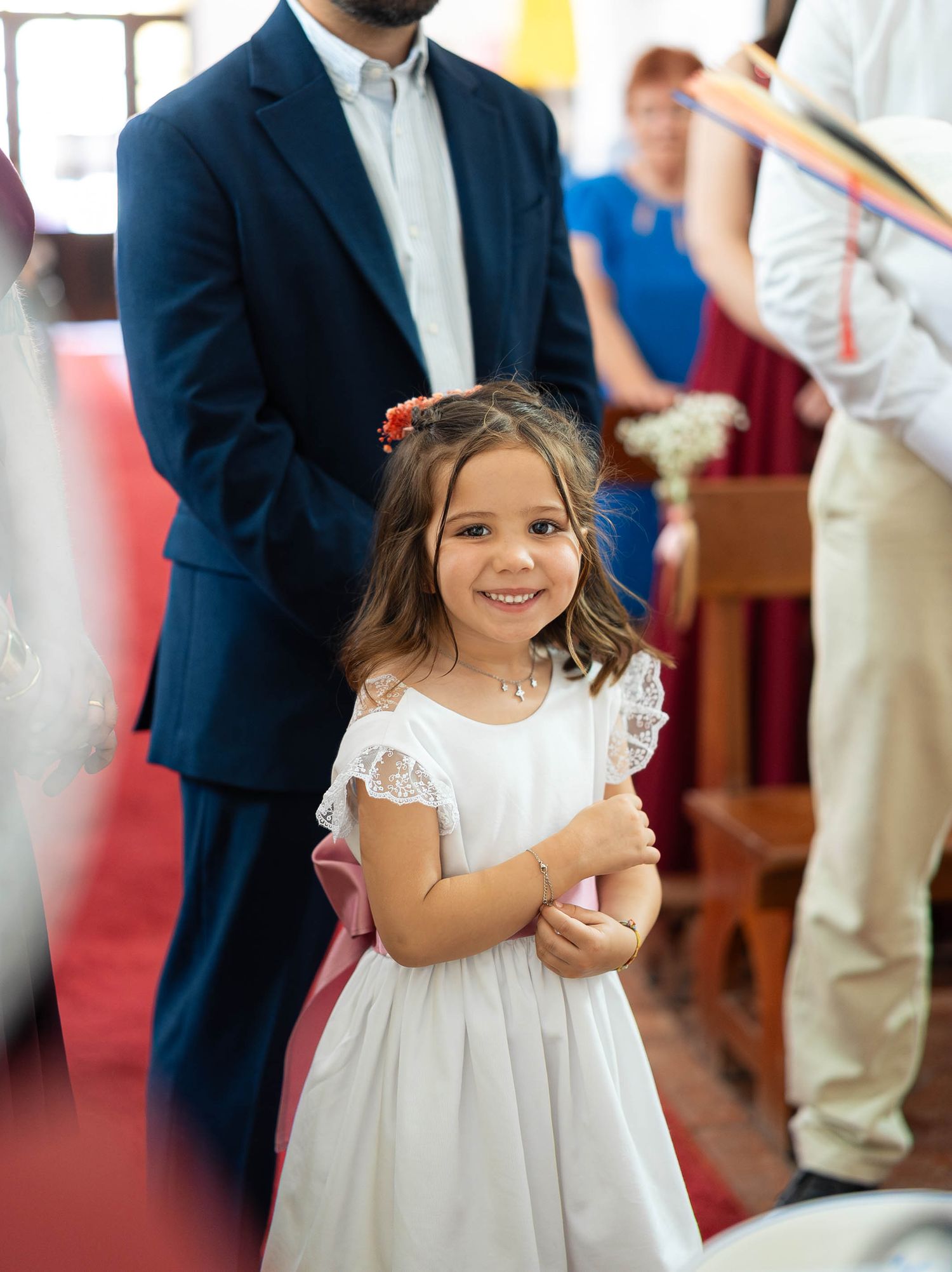 A smiling flower girl in a white dress with pink sash stands in a church aisle during a wedding ceremony.