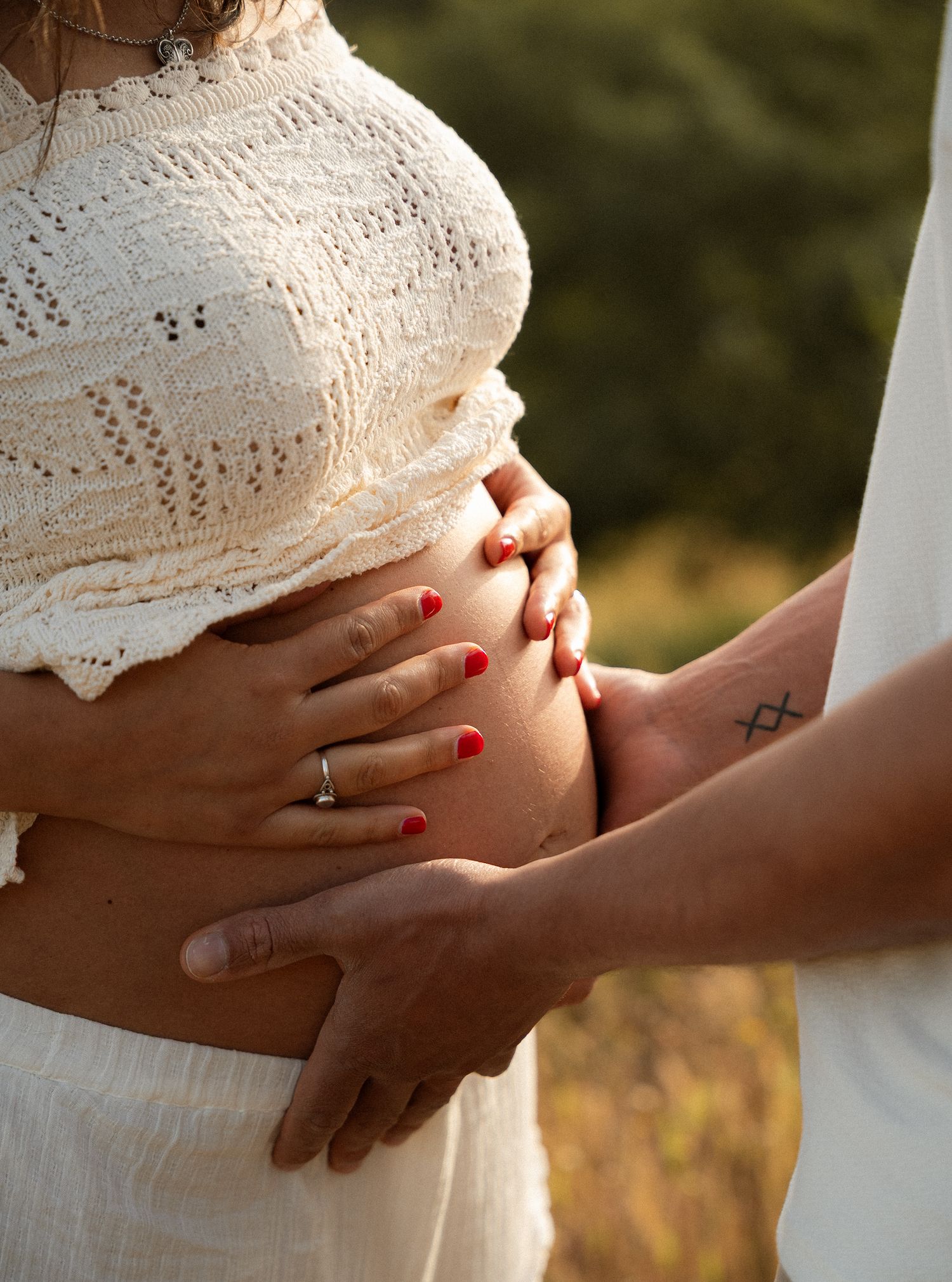 Pregnant woman in white lace crop top cradling baby bump, partner's hand with tattoo resting alongside outdoors.