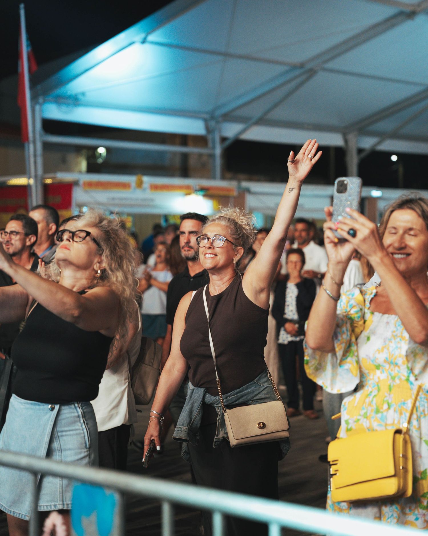 Enthusiastic crowd watching and cheering at an outdoor event under a white tent.