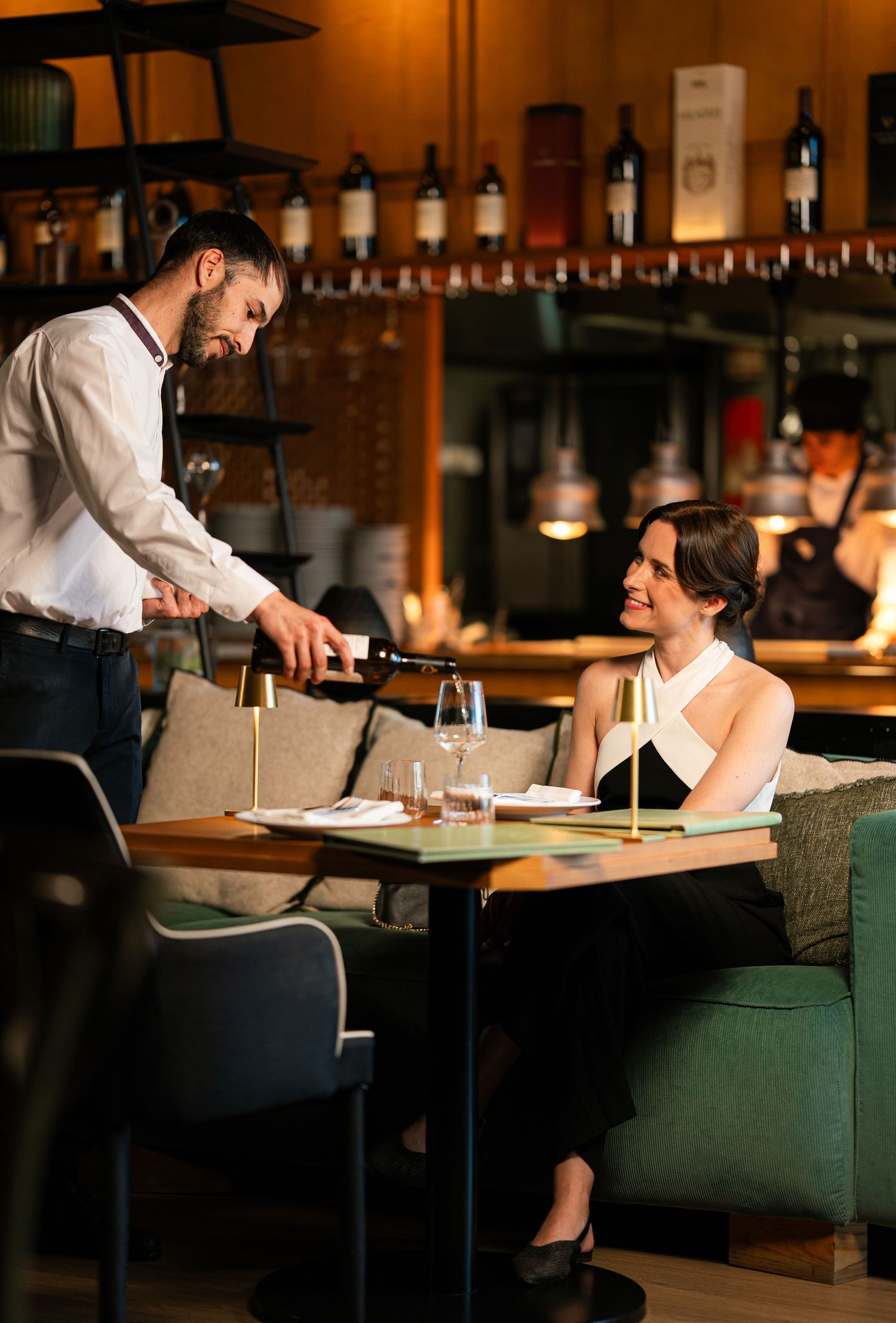 Waiter pouring red wine for a smiling woman in a black dress at an elegant restaurant table.
